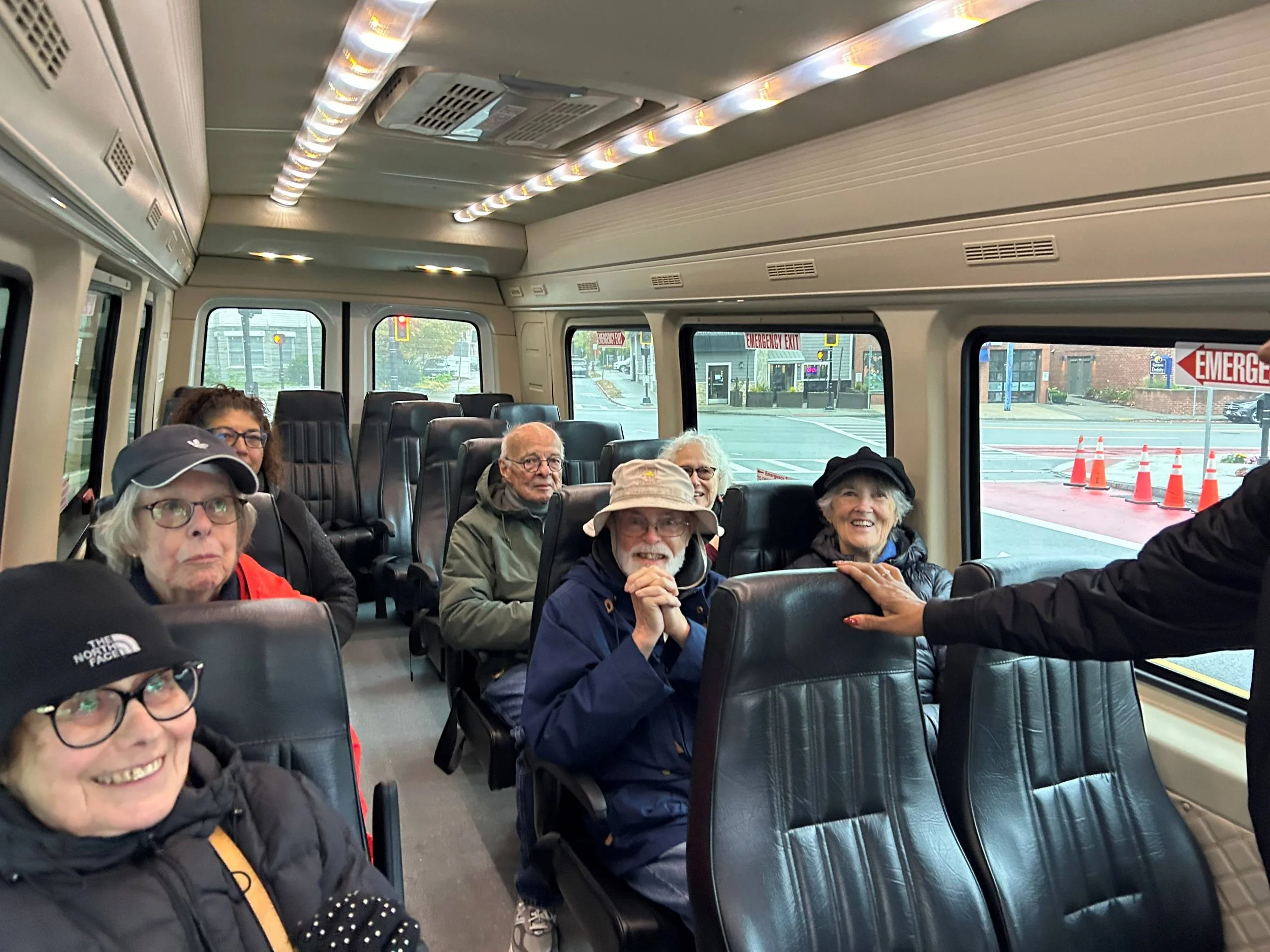Interior of a bus with six passengers seated, some smiling and some looking toward the camera, with large windows showing outside street scene with traffic cones and signs.