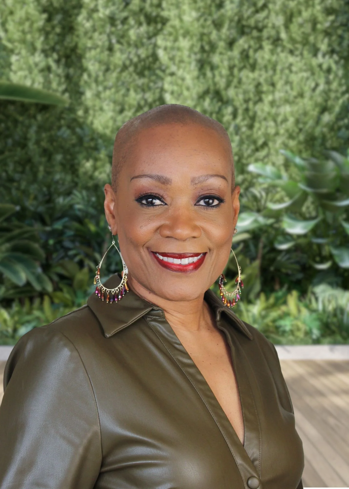 A photo of the Director of WTMA, wearing a dark green, shiny leather blouse and colorful hoop earrings, smiling with a background of green foliage.