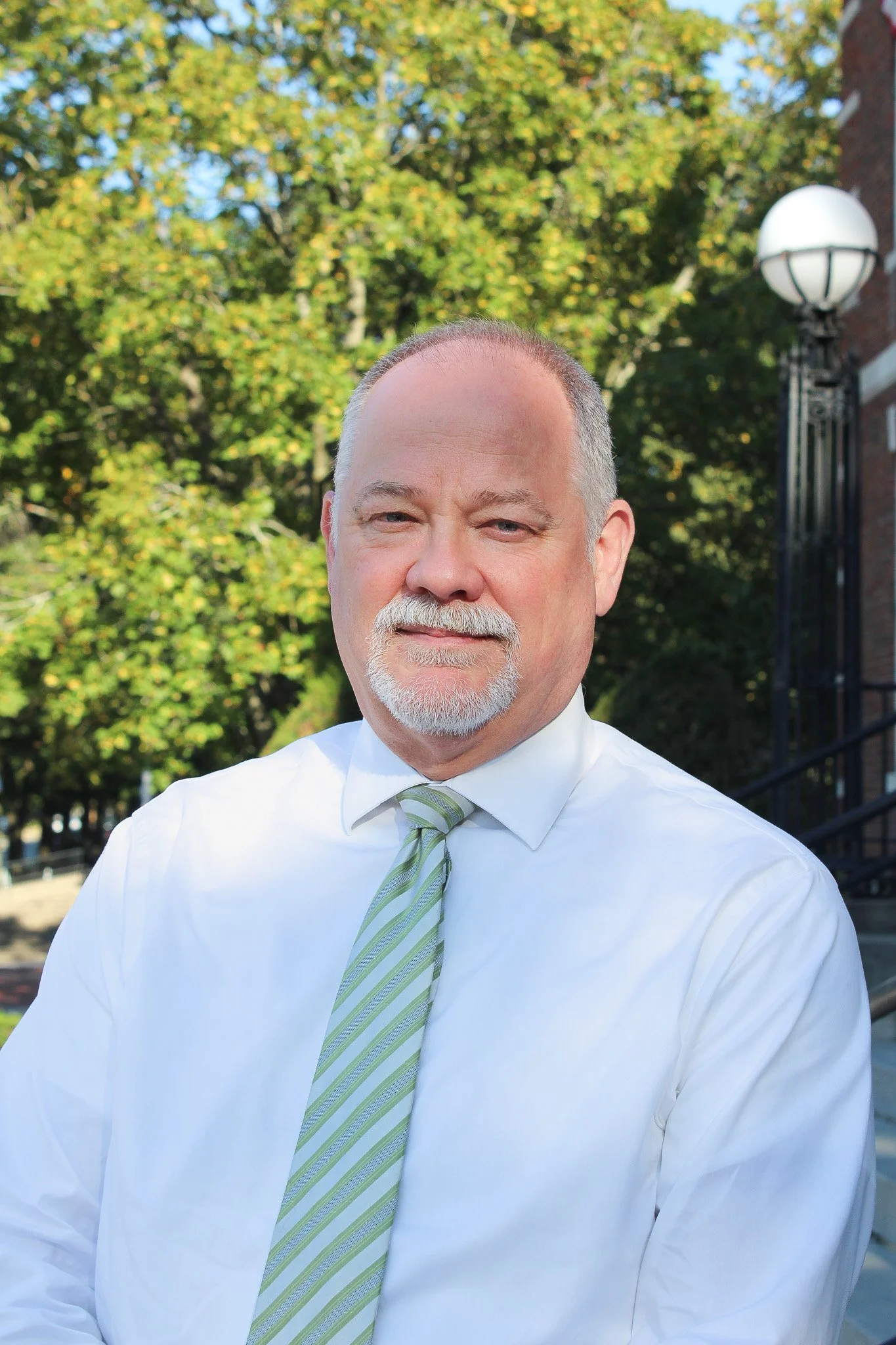 A middle-aged man with a beard and mustache wearing a white shirt and a light green striped tie standing outdoors with trees and a brick building in the background.
