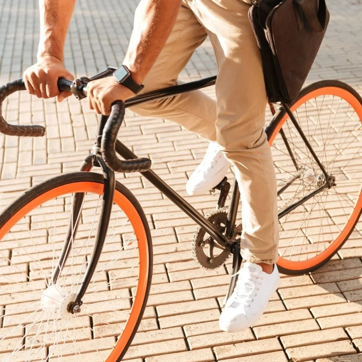 A person riding a black bicycle with orange wheels on a brick pavement, wearing beige pants, white sneakers, and a black watch, with a black bag clipped on the bike's frame.