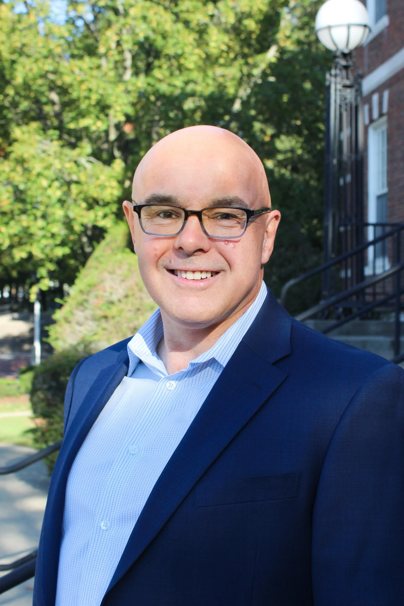 A man with glasses in a blue suit and light blue shirt standing outdoors on a sunny day, smiling with trees and a brick building in the background.