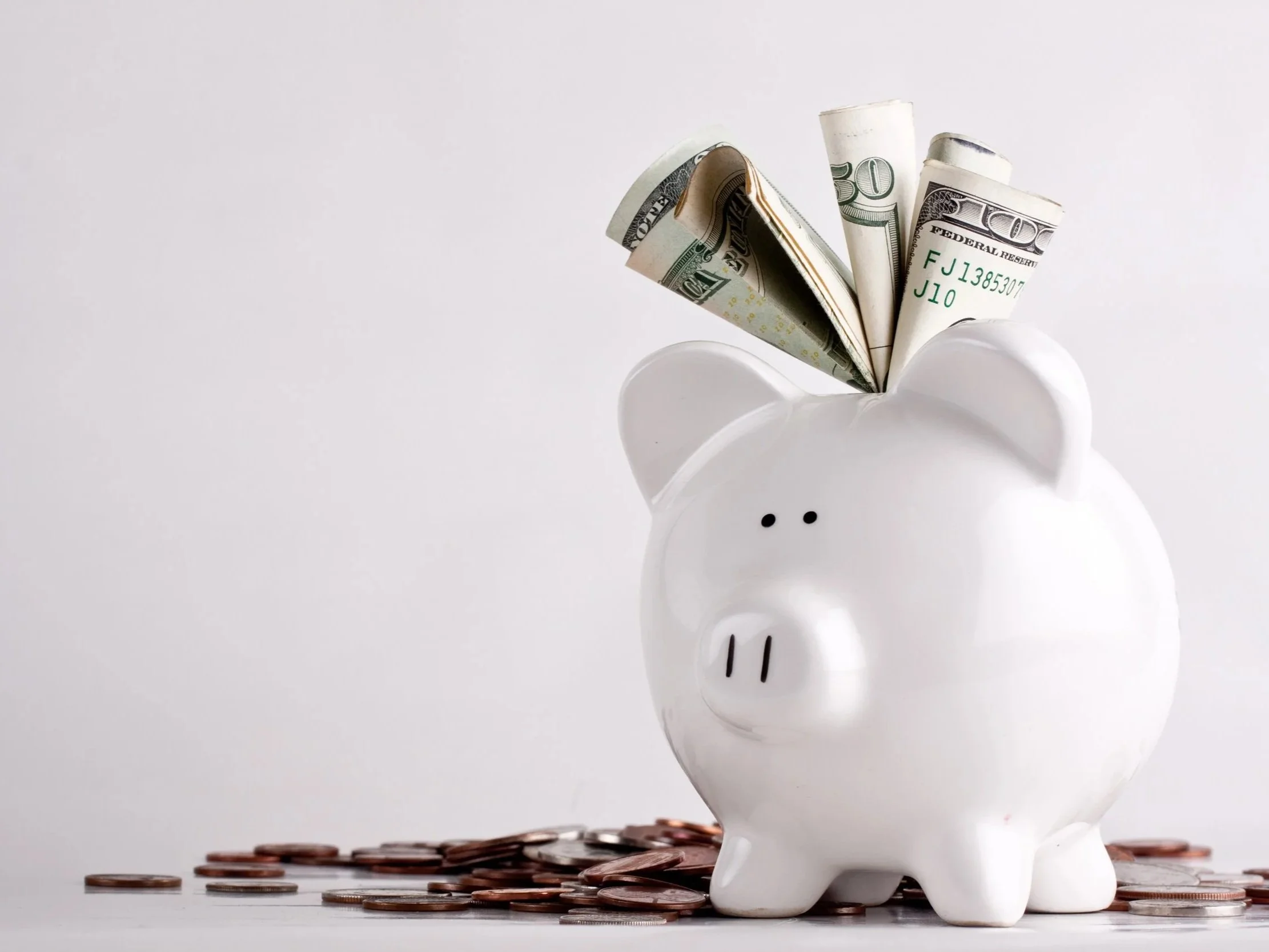 A white piggy bank with rolled dollar bills and scattered coins on a white surface.