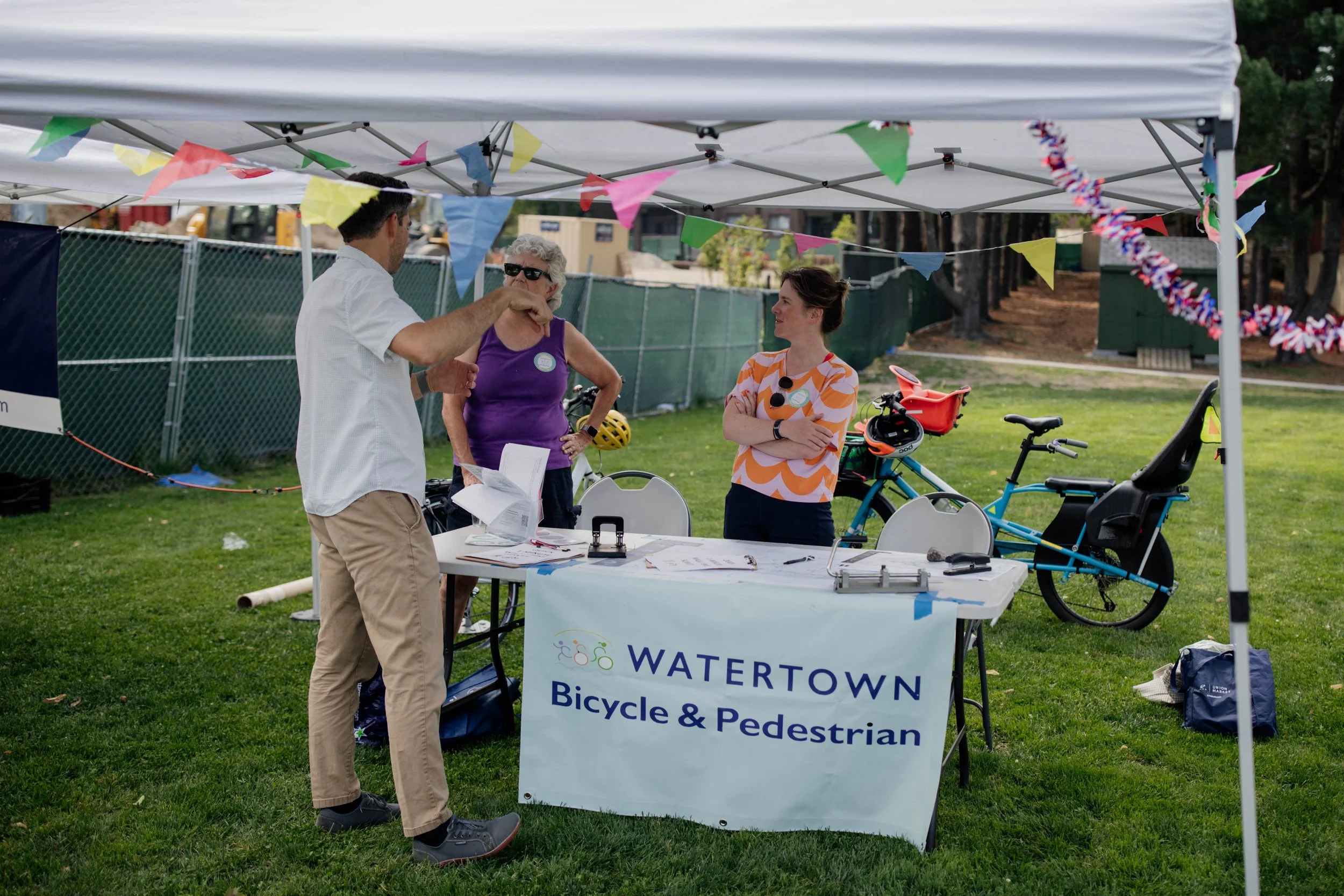 A small outdoor booth with a banner that reads "WATERTOWN Bicycle & Pedestrian". Two women and one man are standing inside the booth, which is decorated with colorful pennant banners. Bicycles and helmets are visible behind the women, and the man appears to be talking or explaining something to the women.