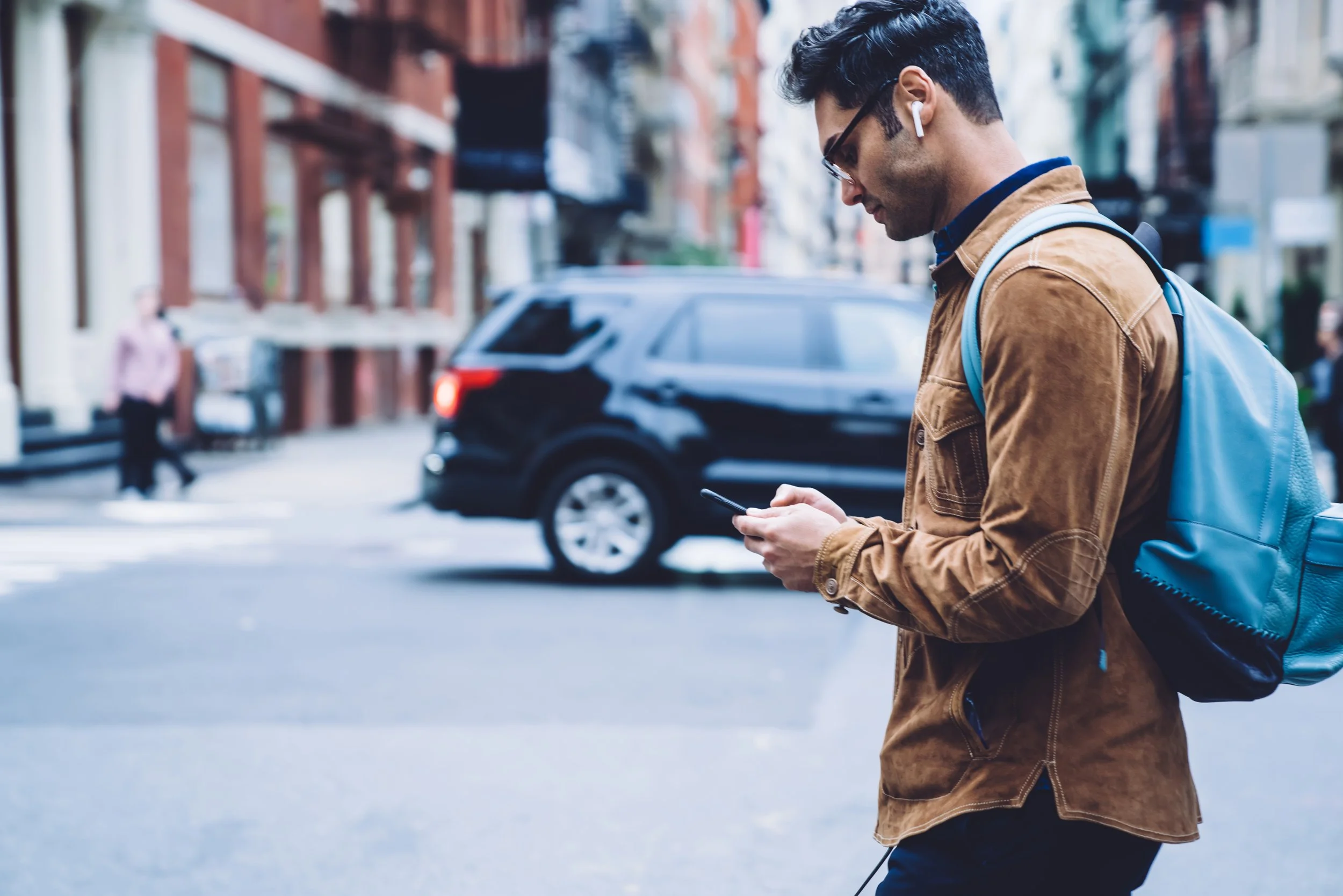 A man with dark hair, sunglasses, and wireless earbuds stands on a city street, looking at his phone while wearing a brown jacket and carrying a blue backpack.