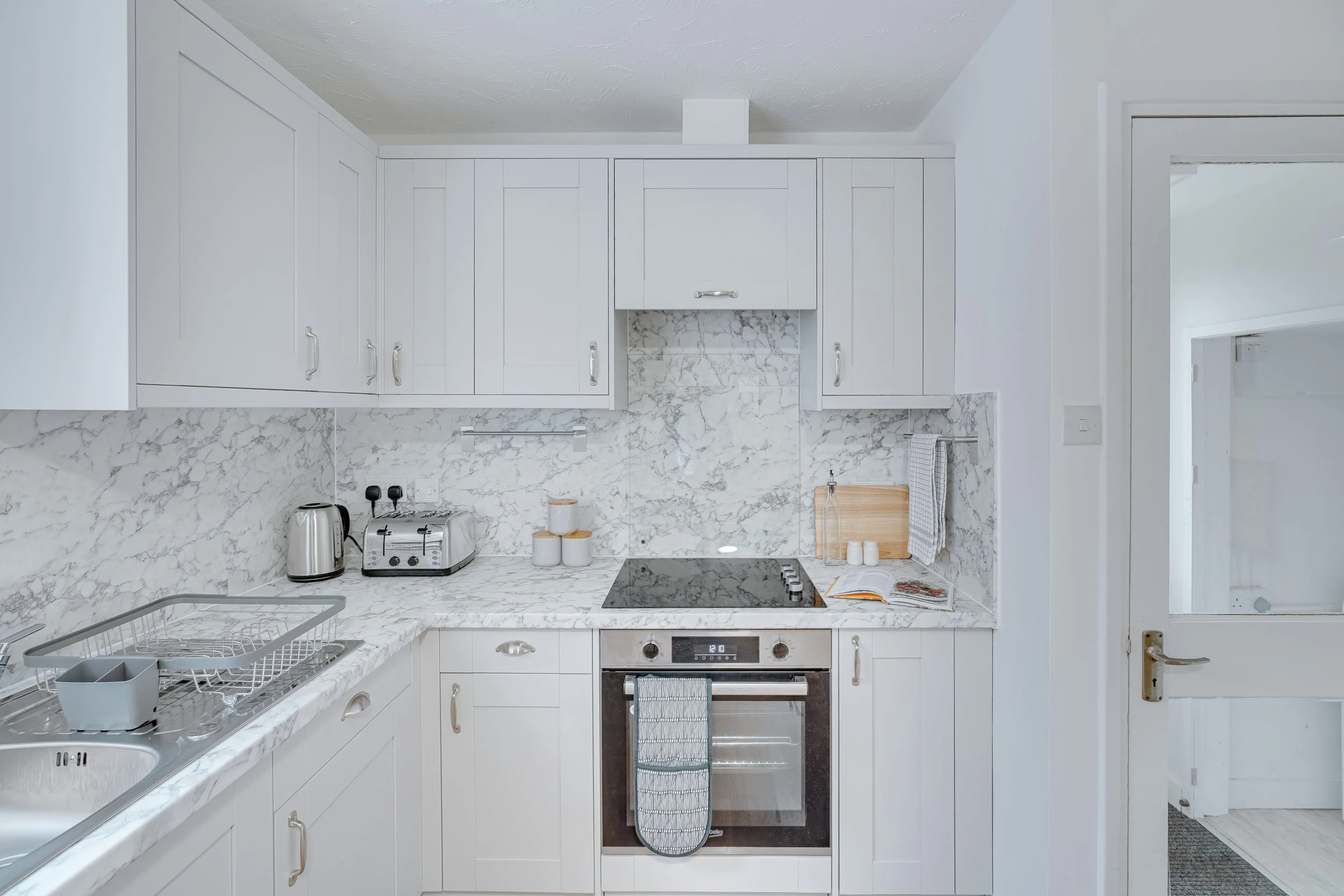 Kitchen with white cabinets, marble backsplash and countertop, stainless steel appliances including a stove and toaster, and a sink with a dish drying rack.