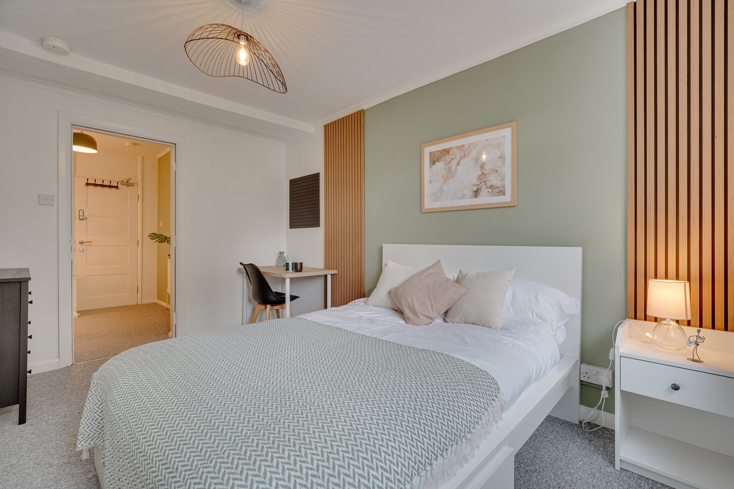 A modern bedroom with a white bed, light green accent wall, and wooden slat decor. A black chair and small wooden desk are in the corner. A nightstand with a lamp is next to the bed, and framed artwork hangs above.