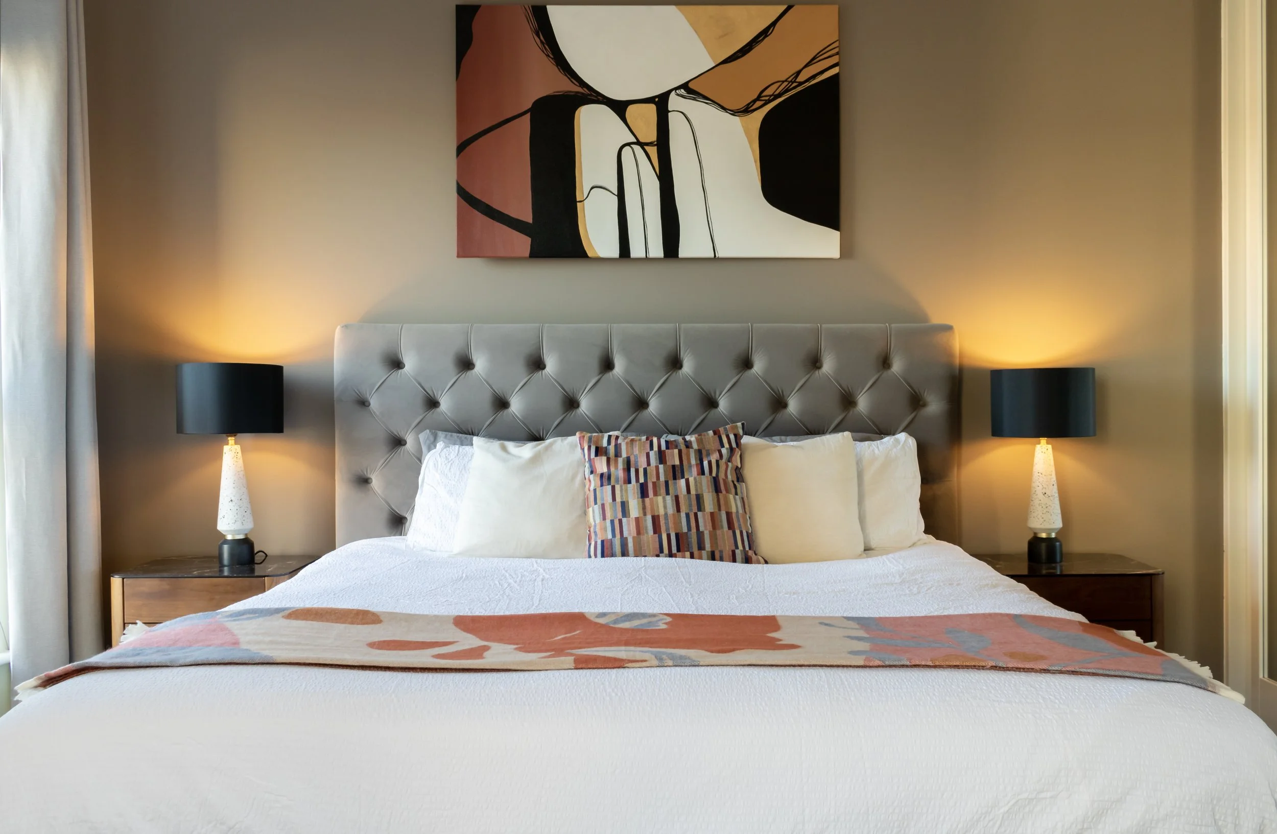 Residential bedroom interior featuring a tufted upholstered headboard, layered neutral bedding, bedside tables with warm ambient lighting, and abstract artwork creating a balanced, inviting space.