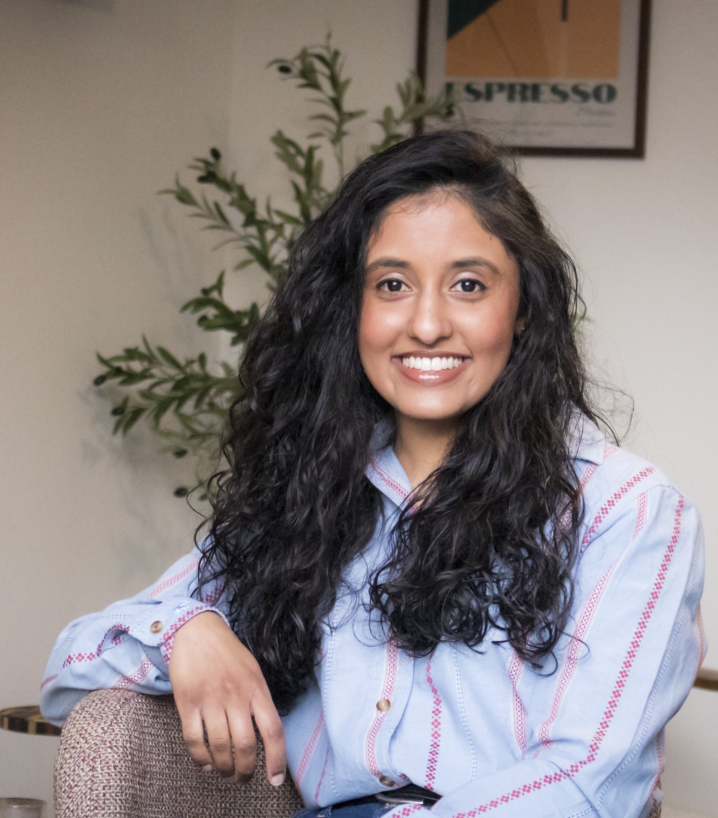 A woman with long, curly dark hair smiling and sitting on a beige chair in a room with a plant and framed artwork in the background.