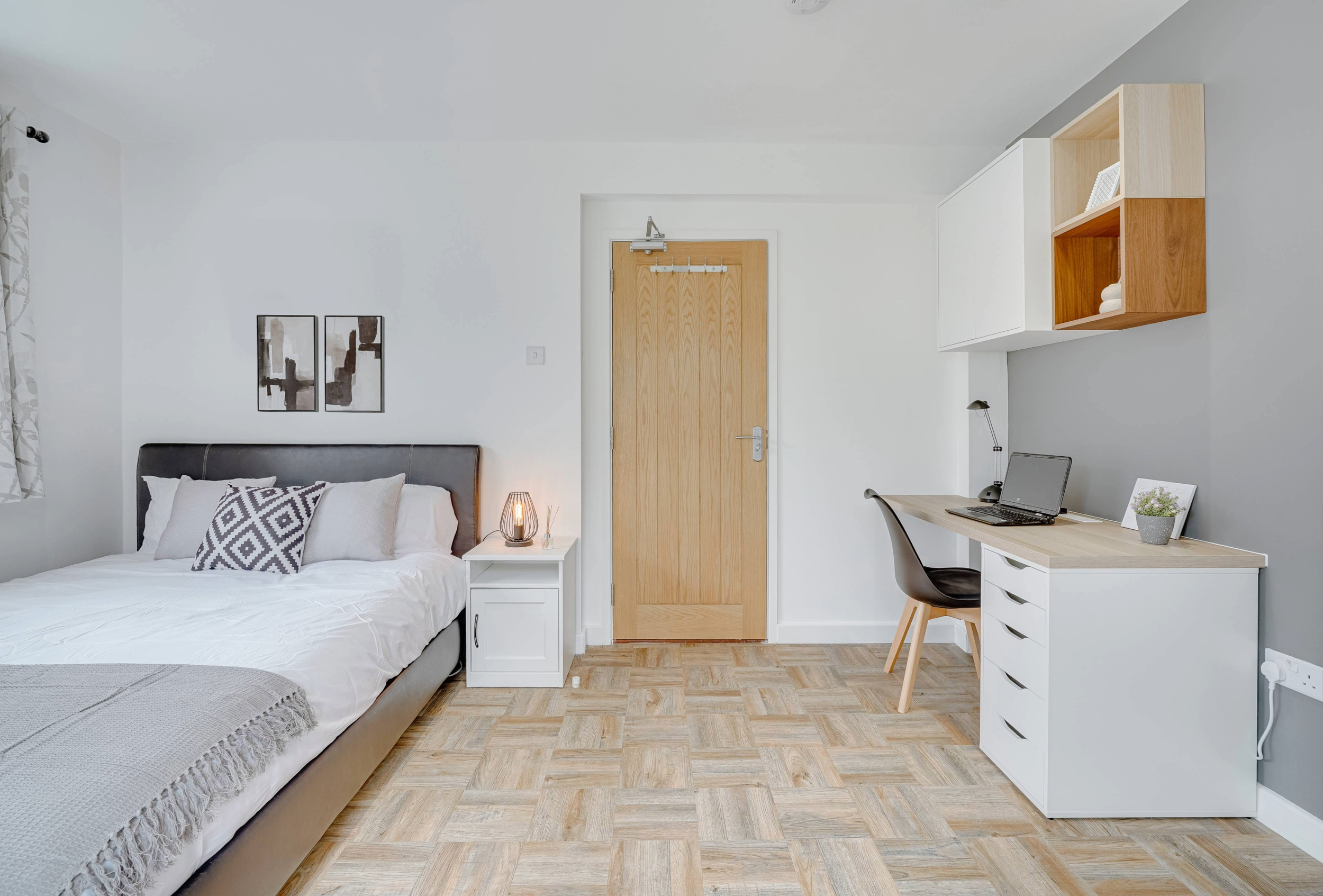 Residential bedroom interior featuring a modern upholstered bed, neutral bedding, bedside table, and an integrated desk workspace with wall-mounted storage in a light, calm colour palette.