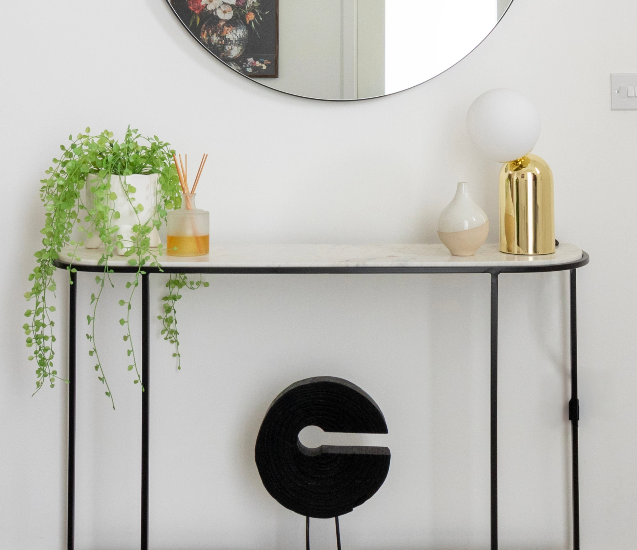 Residential interior styling detail featuring a slim console table styled with decorative objects, greenery, a table lamp, and a round mirror, creating a refined and welcoming entrance space.