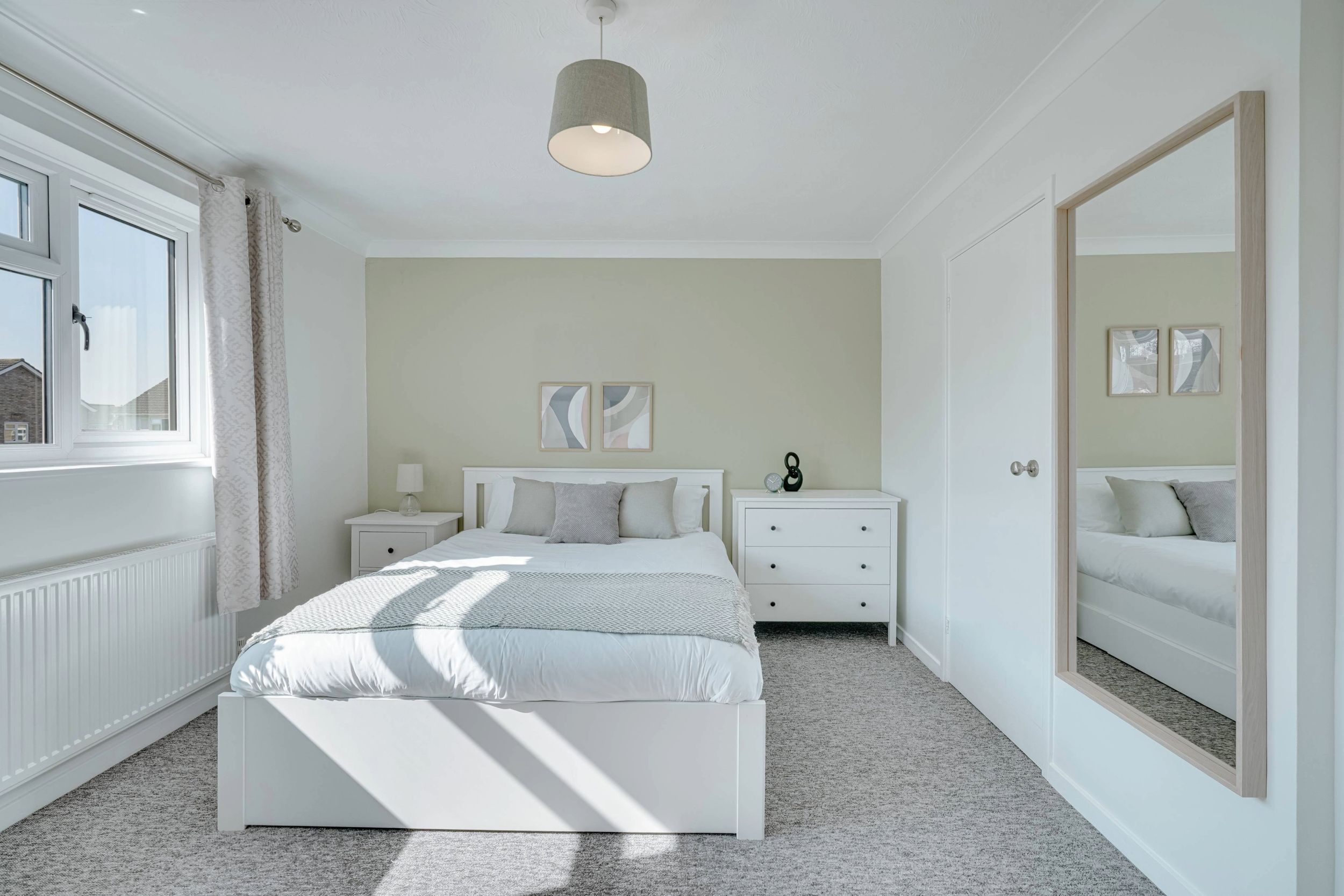 A bright, modern bedroom with a white bed, grey and beige decor, a large window with curtains, and a mirror reflecting the room.