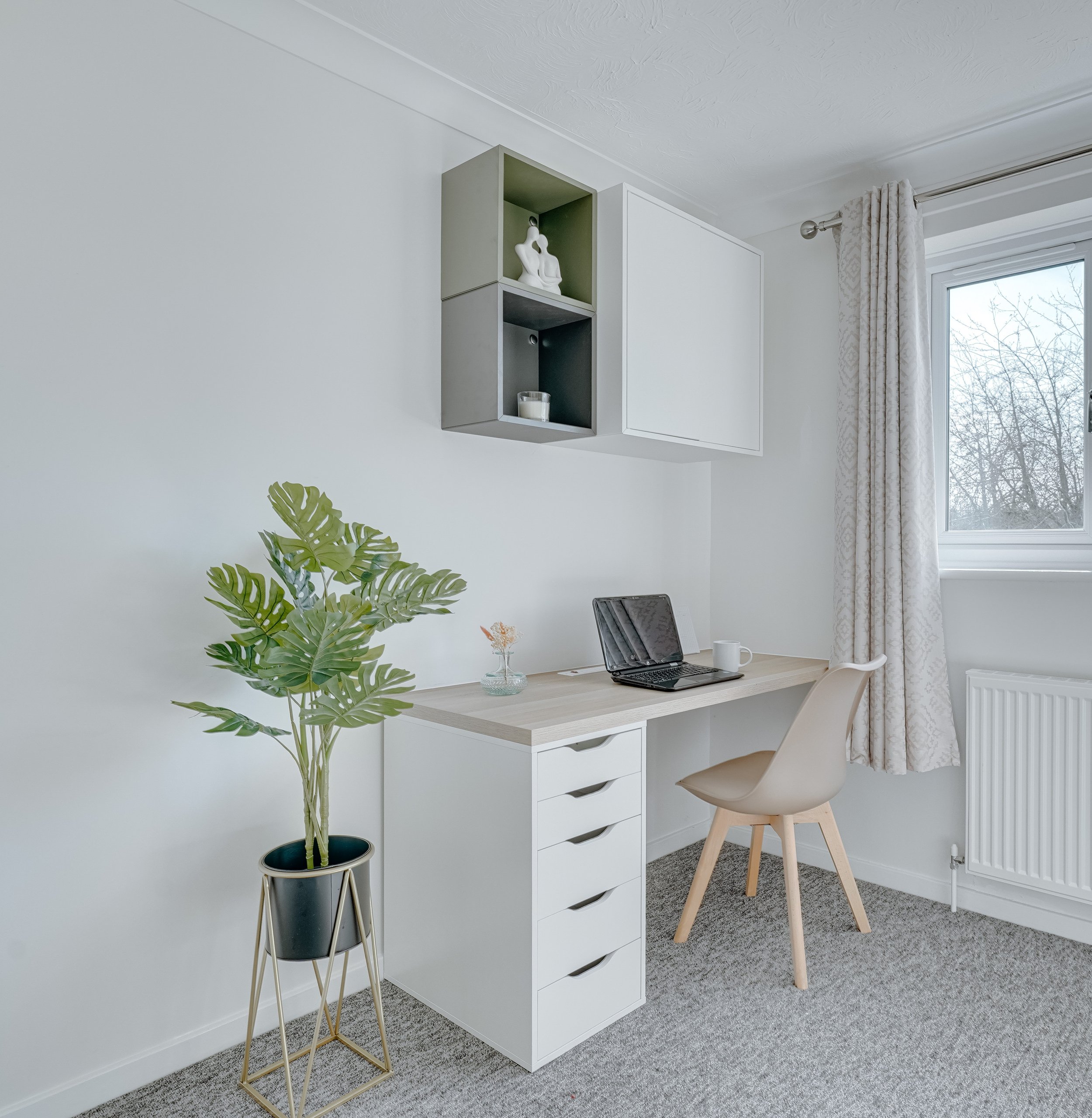 Residential bedroom interior featuring a compact desk workspace with integrated drawers, wall-mounted storage, a simple chair, and soft natural light creating a clean, functional setting.