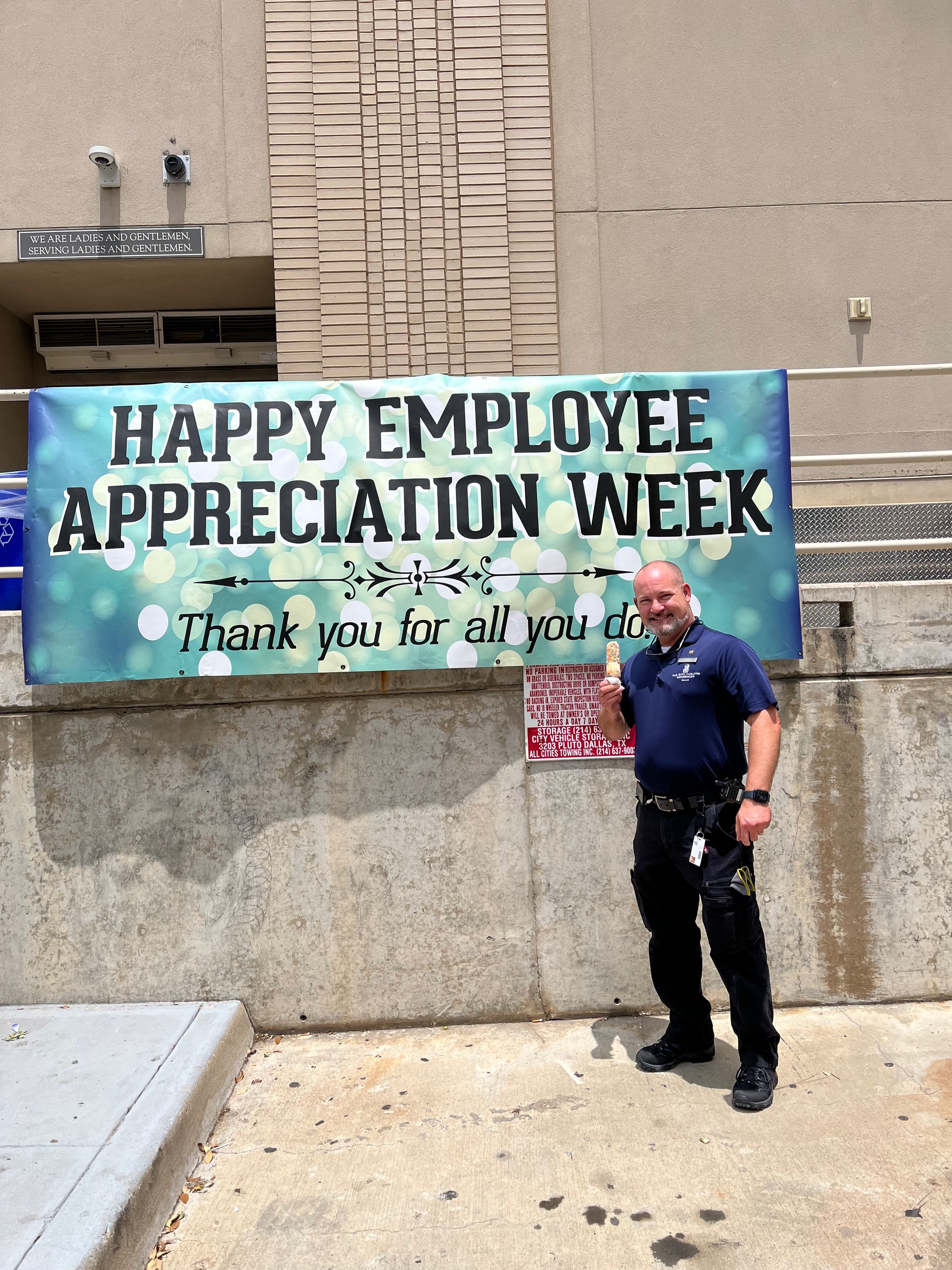 A man in uniform standing in front of a large banner that reads 'Happy Employee Appreciation Week. Thank you for all you do'. The man is smiling and holding an ice cream bar.