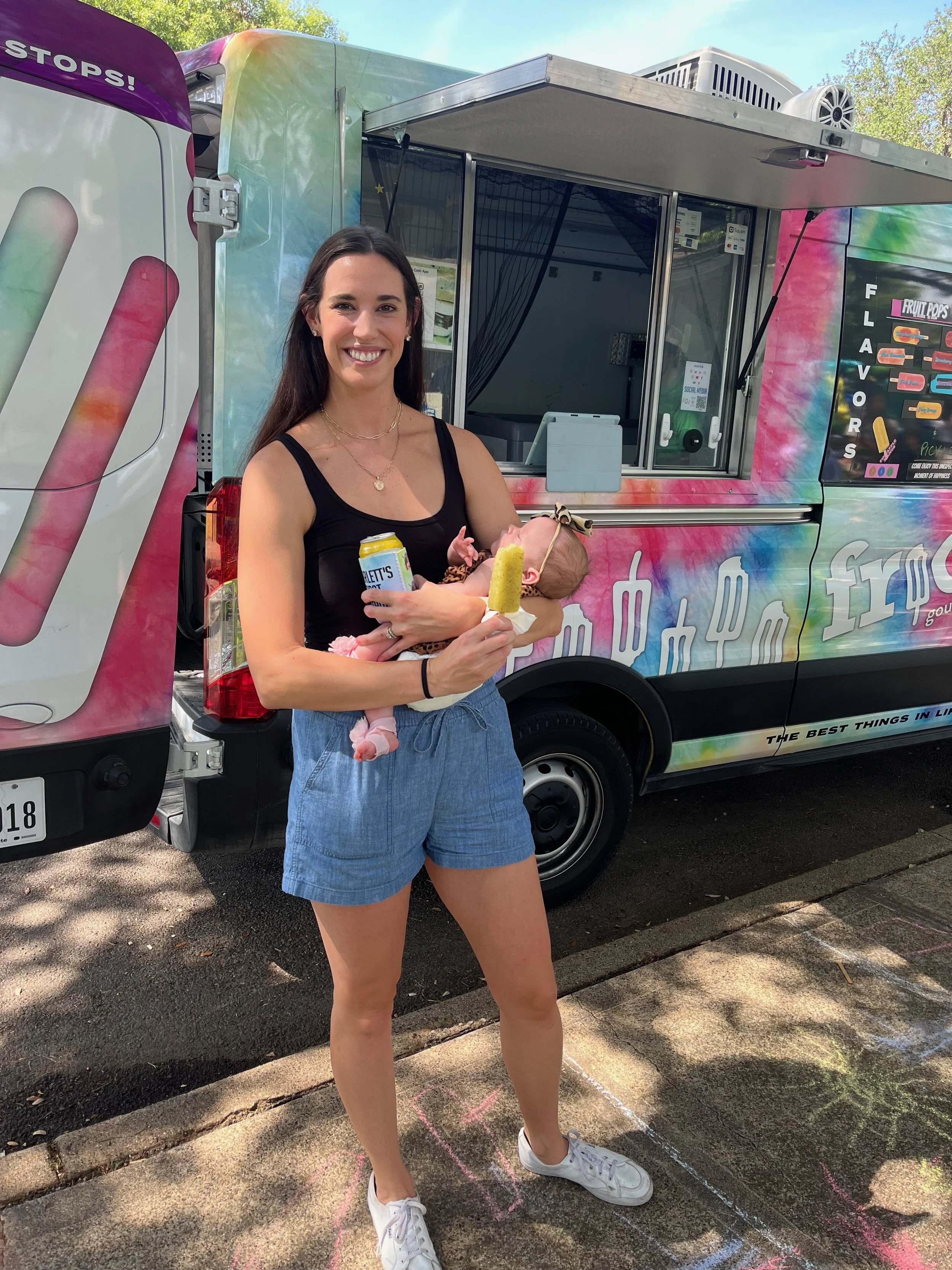 Woman smiling and holding a baby and a yellow popsicle in front of a colorful ice cream truck.