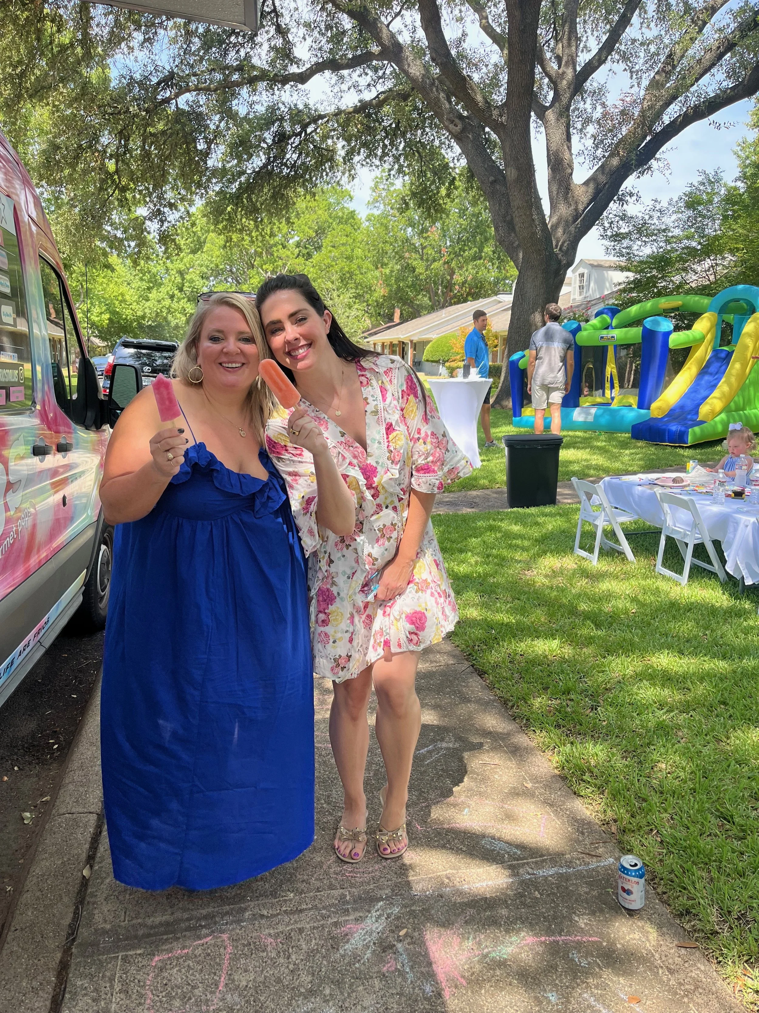 Two women smiling and holding popsicles at an outdoor birthday party, with a bounce house and party tables in the background under a large tree.