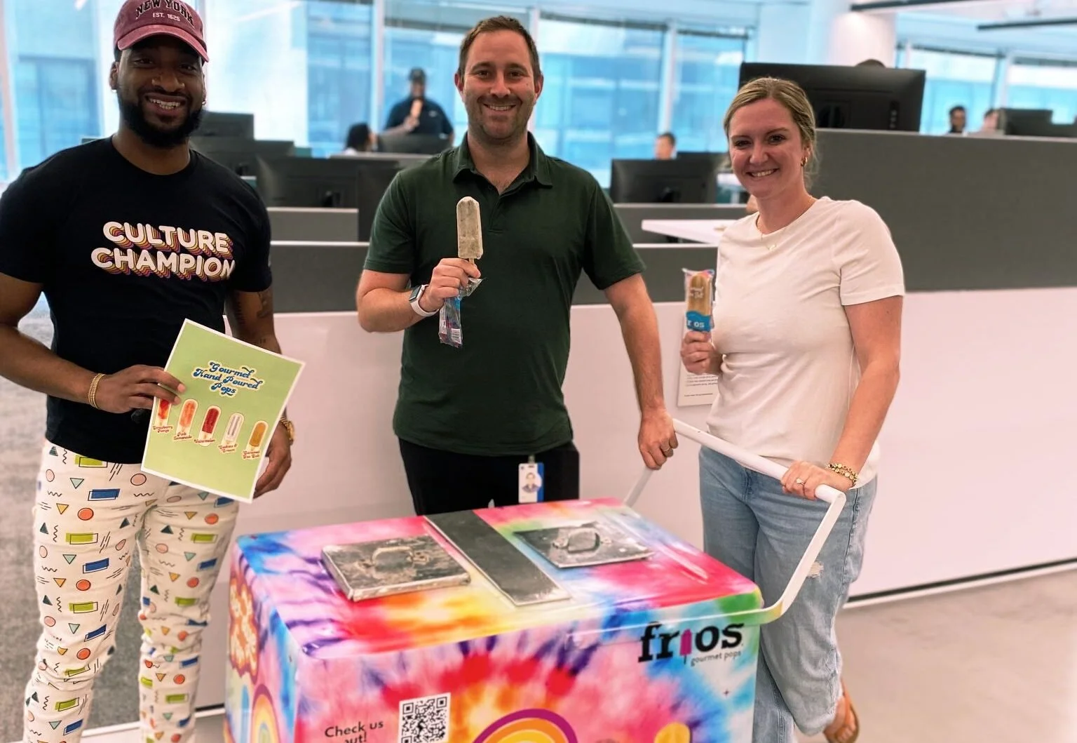 Three people are standing behind a table with colorful fabric cover, at an indoor event space. They are holding ice cream bars and smiling at the camera. The person on the left is wearing a black T-shirt and patterned pants, holding a sign. The person in the middle is wearing a green polo shirt, holding an ice cream. The person on the right is wearing a white T-shirt and jeans, also holding an ice cream.