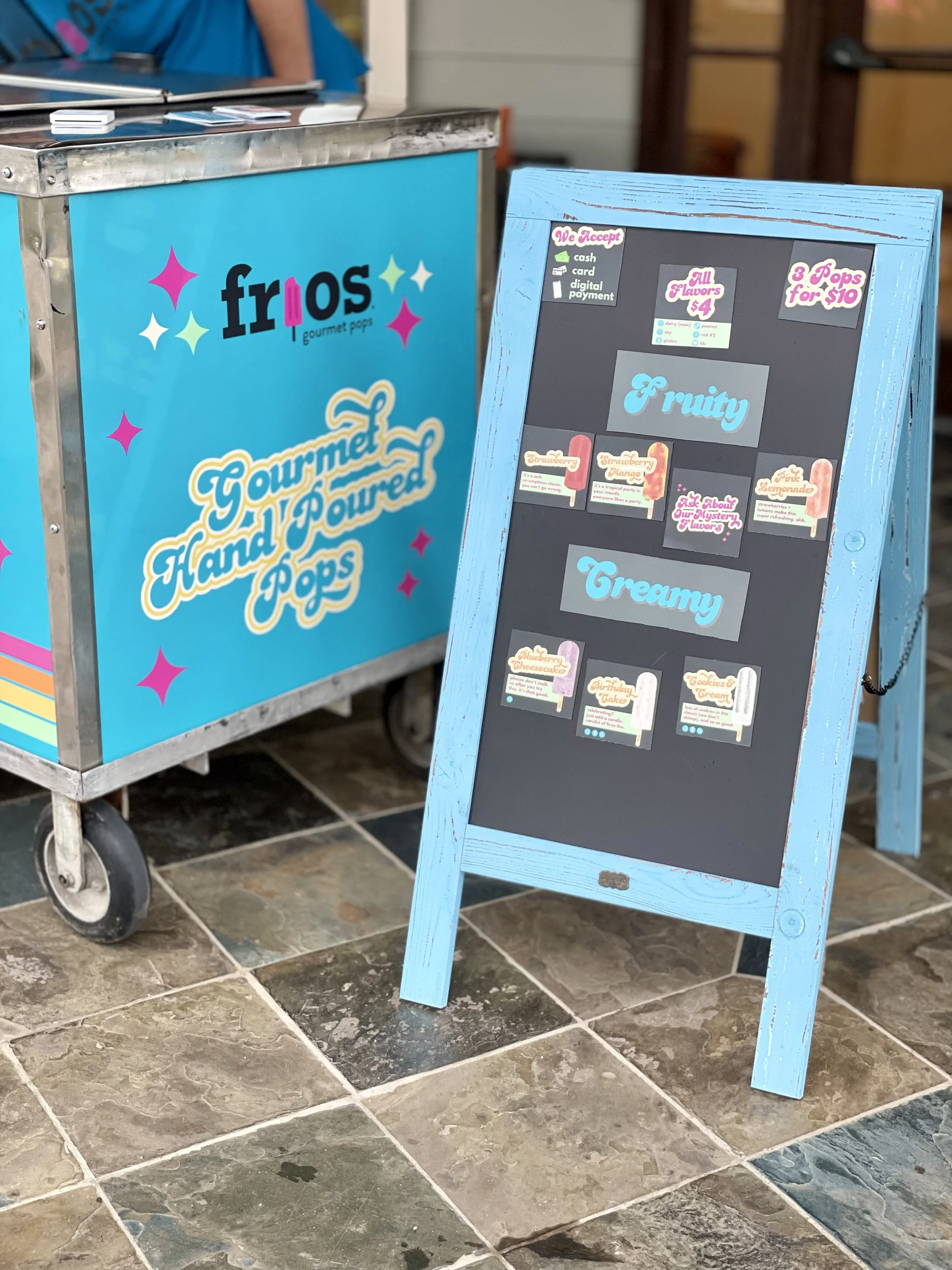 A blackboard-style menu sign and pop cart of Frios Dallas. The menu offers different flavored Frios Pops