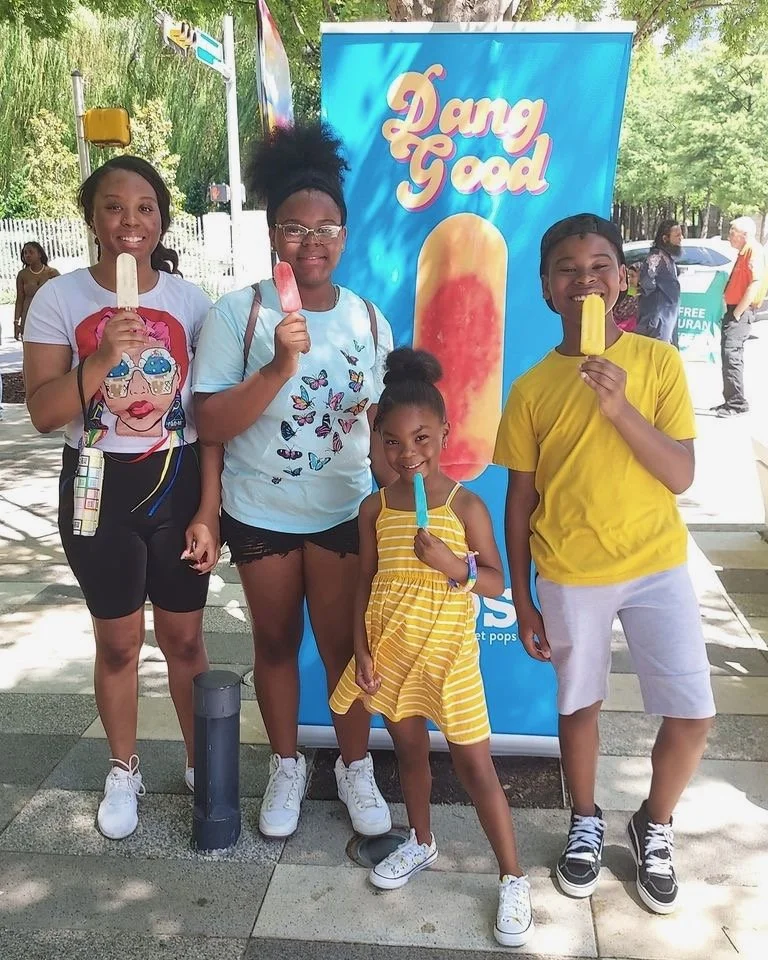 Four children standing outdoors, each holding a colorful ice pop, posing in front of a bright blue sign that says 'Dare Good,' with trees and people in the background.