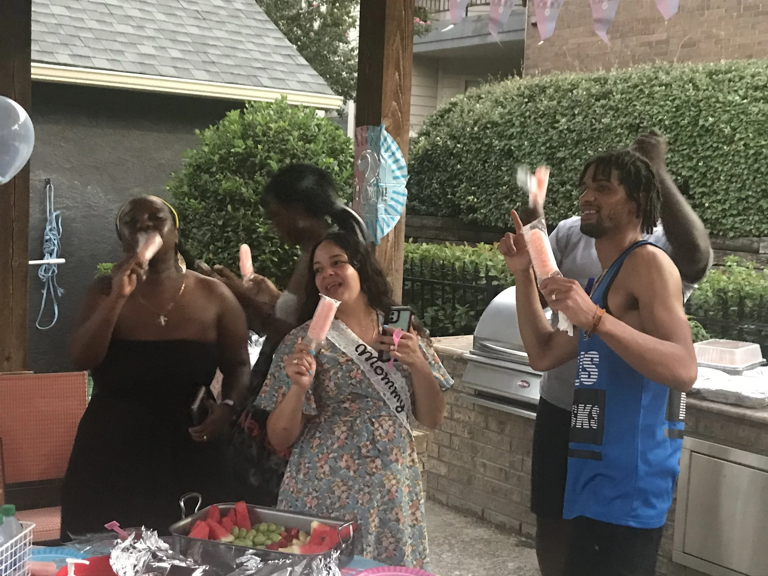 A diverse group of people celebrating at a backyard party, enjoying watermelon and holding pink popsicles, with decorations including balloons and banners.
