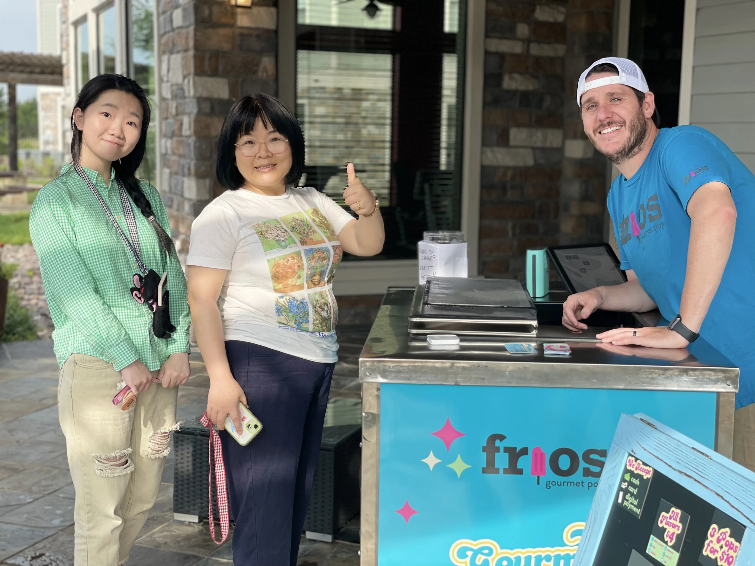 Three people standing at an outdoor Frios cart stand, smiling at the camera. The stand has a vendor, with a menu board in front.