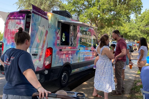 People lining up at a colorful ice cream truck in a park, with trees and parked cars in the background.