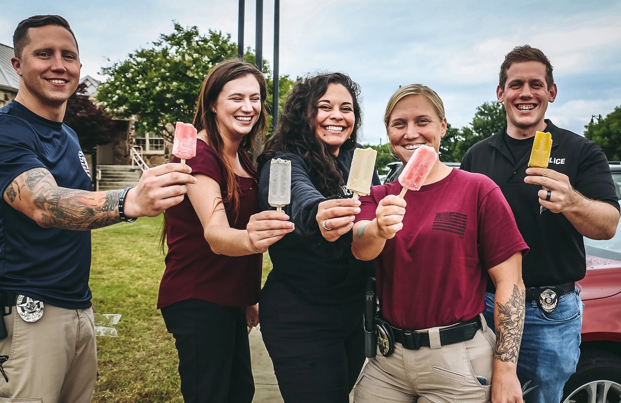 Group of five smiling police officers holding colorful popsicles outdoors on a cloudy day.