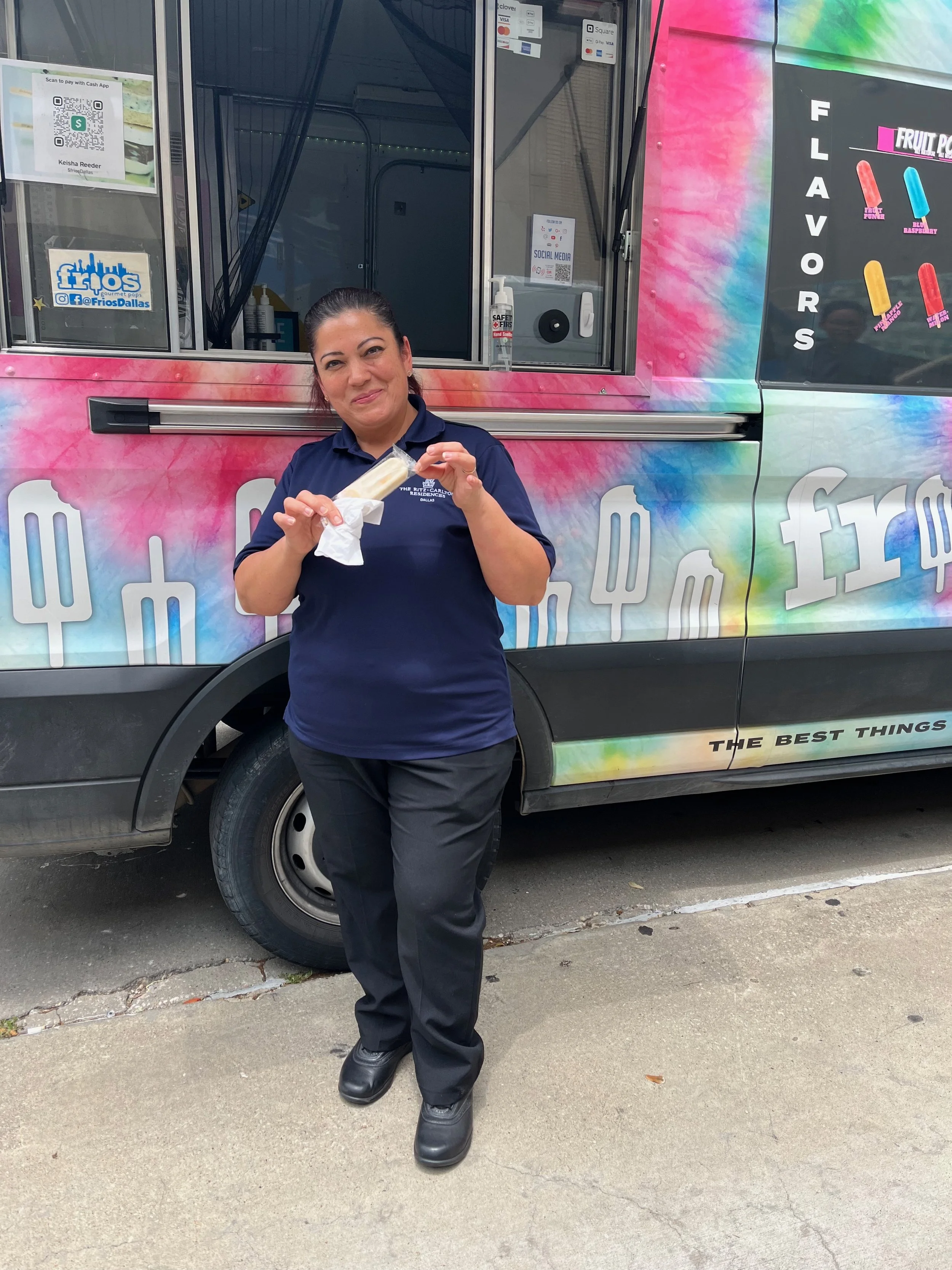 A woman in a navy blue uniform standing in front of a colorful ice cream truck, holding a vanilla ice cream cone with a napkin, smiling at the camera.