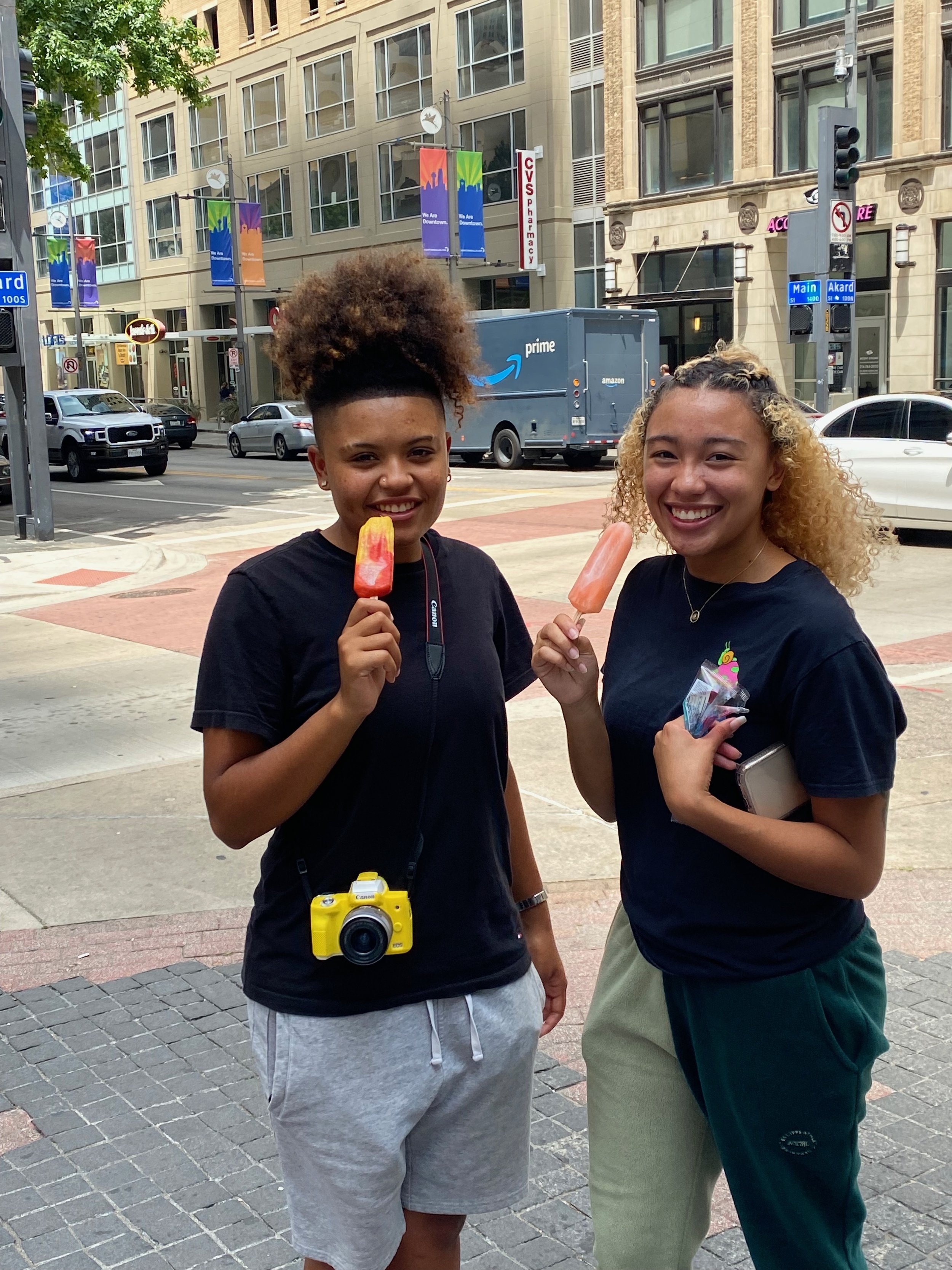 Two smiling women holding popsicles and standing on a city sidewalk with buildings, cars, and traffic lights in the background.