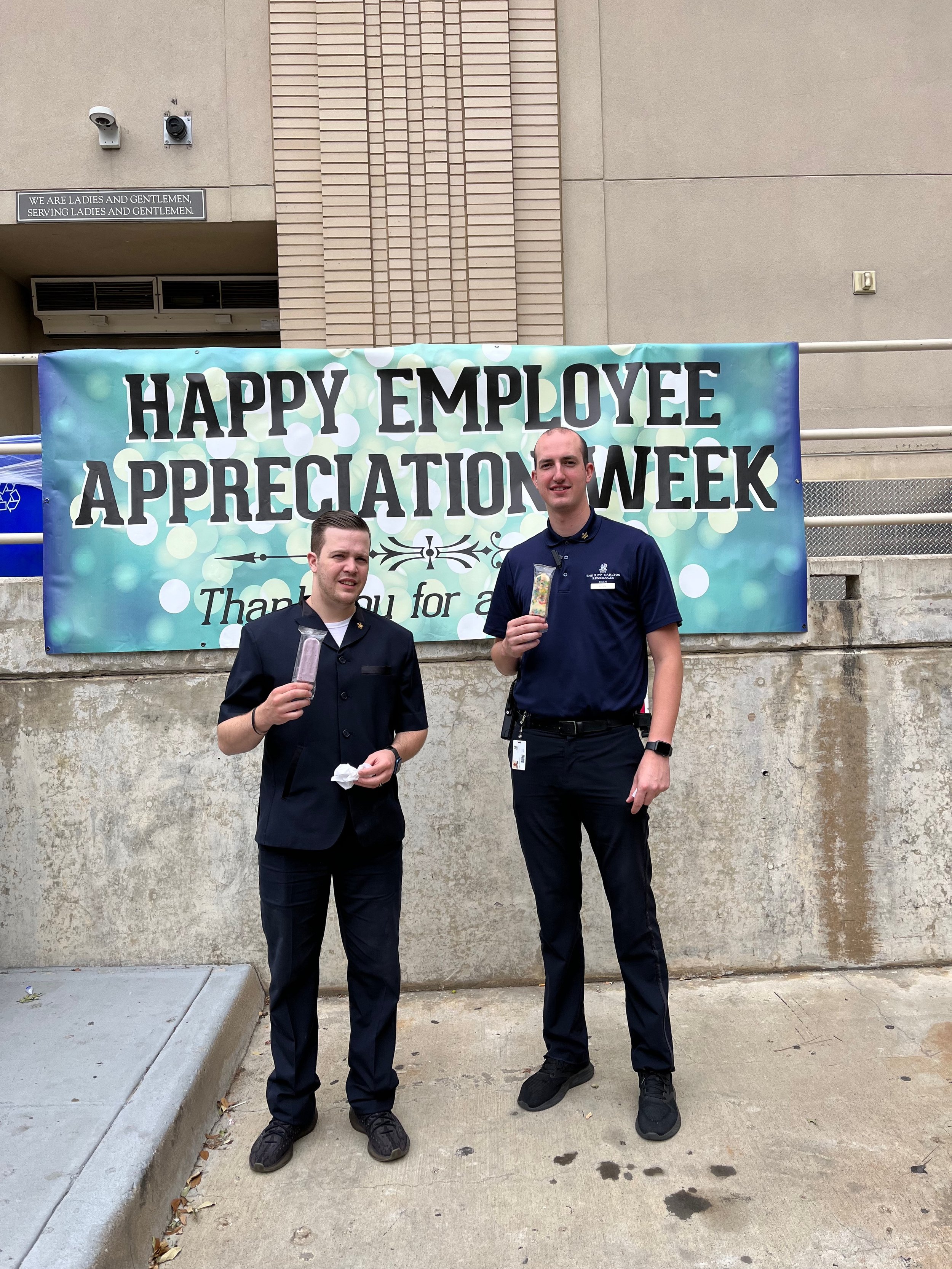 Two employees standing in front of a banner that reads 'Happy Employee Appreciation Week,' holding ice cream treats, at a workplace celebration.