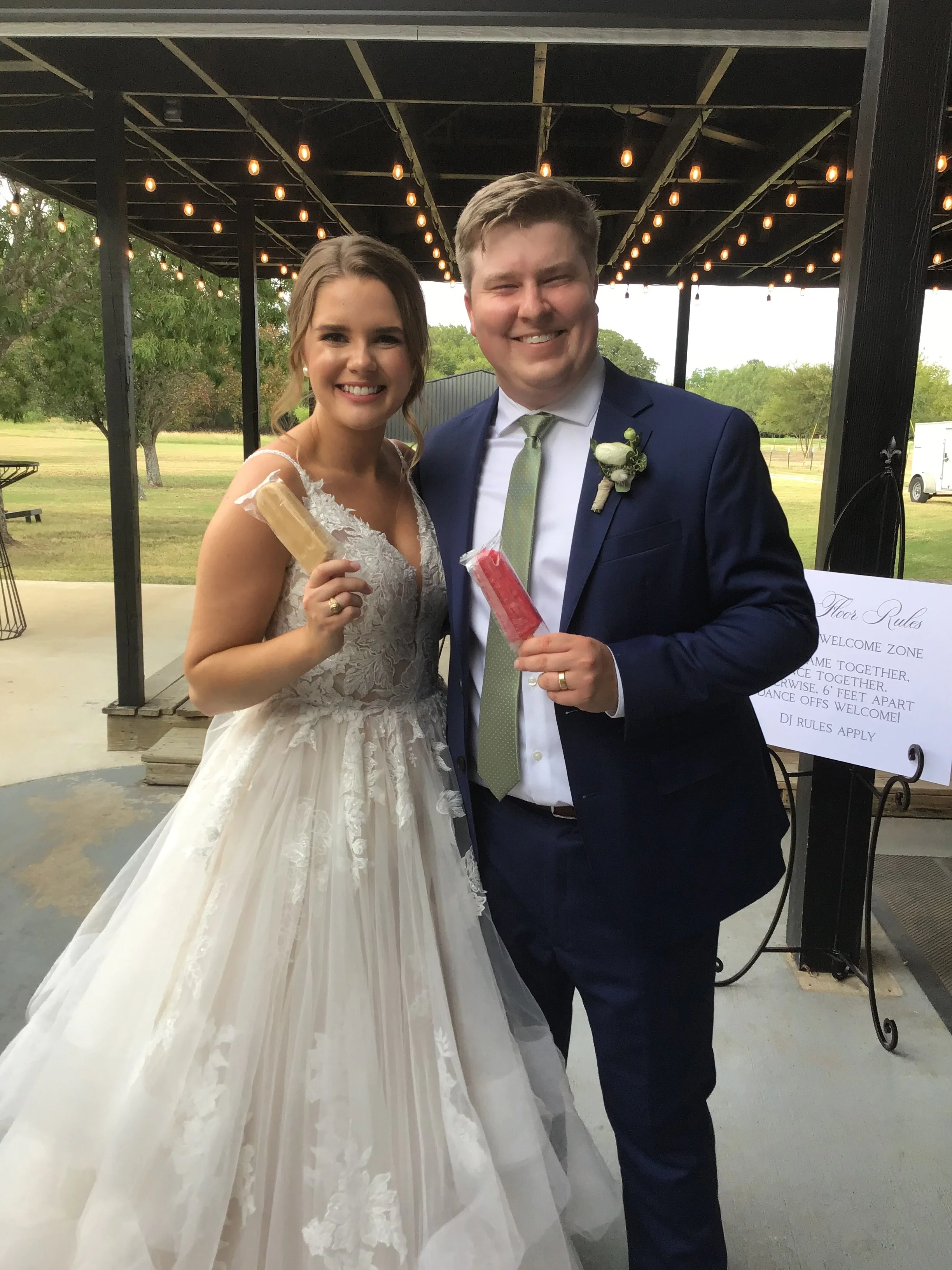 Bride and groom smiling at a wedding reception, holding candy sticks, standing under a decorated outdoor pavilion with string lights.