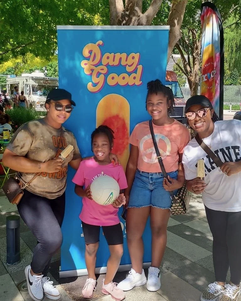 Four women and a girl standing together outdoors, smiling, next to a colorful 
