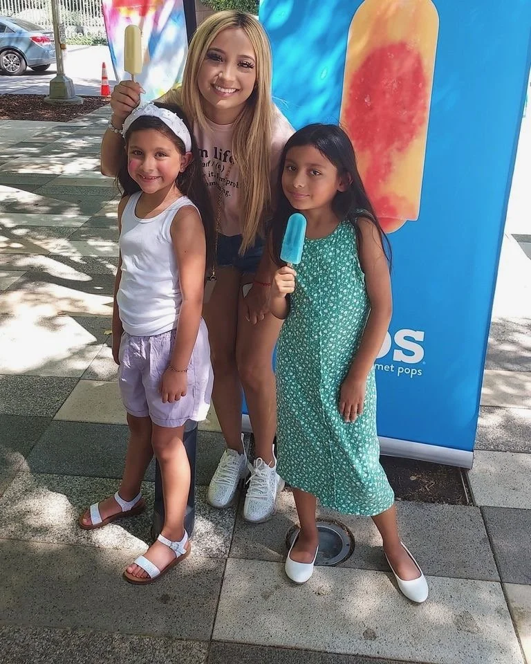 Three females standing outdoors on a sidewalk, smiling at the camera, holding popsicles, with a colorful banner behind them.