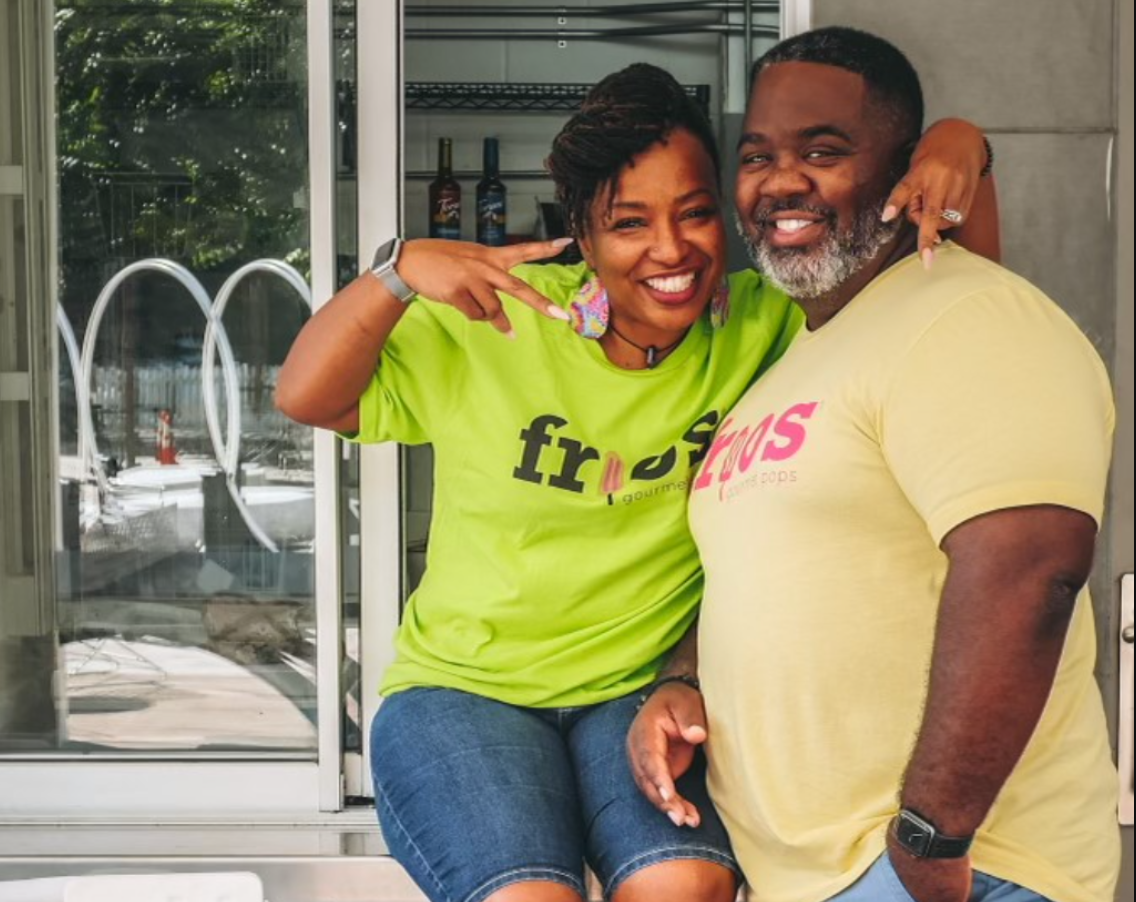 A woman and a man happily pose together, with the woman sitting on a surface and the man standing beside her, smiling at the camera inside a cafe or restaurant.