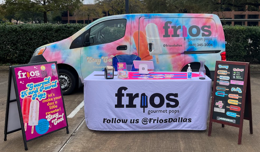 Frios Ice cream truck with colorful tie-dye design and 'frios' branding, set up at an outdoor event with a table and signage promoting gourmet popsicles.
