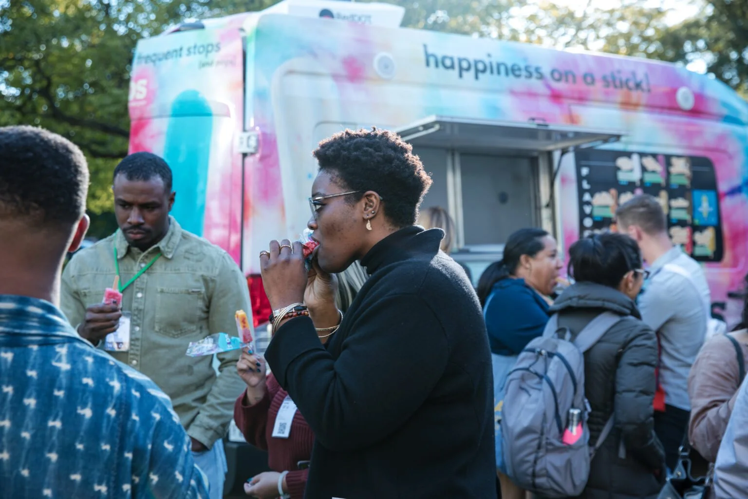 People gathered outdoors around a colorful ice cream truck, some enjoying ice cream and others waiting in line.