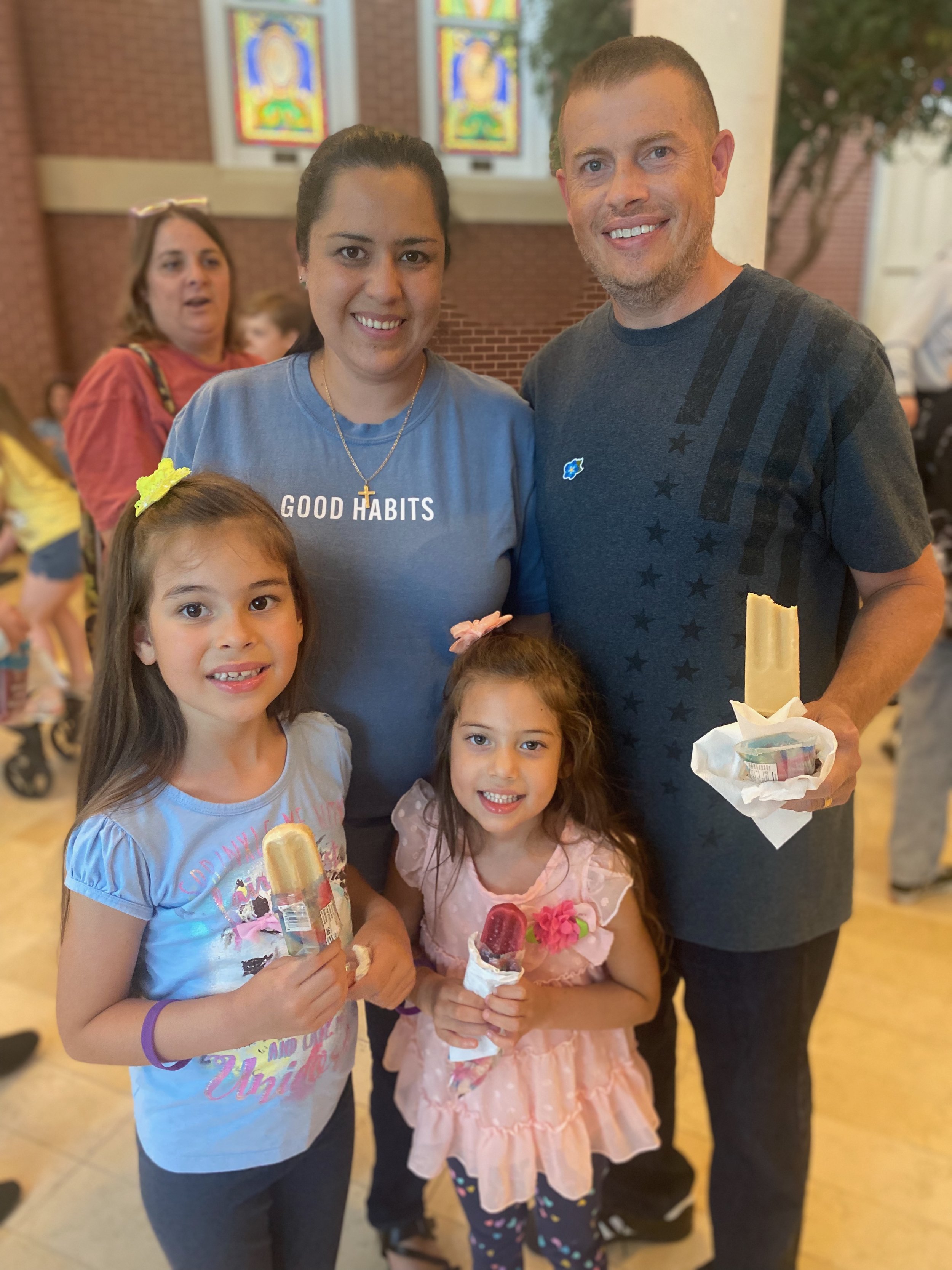 A family of four at an indoor event, two young girls holding ice cream and the parents standing behind them, with stained glass windows in the background.