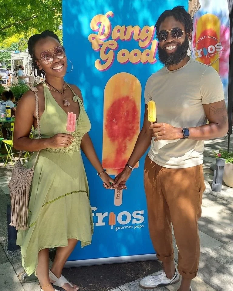 A smiling couple holding hands in front of a colorful ice cream banner with the words 'Dang Good' and 'Trios gourmet pops'. They are holding yellow and pink popsicles, and are both wearing sunglasses and casual summer clothing.