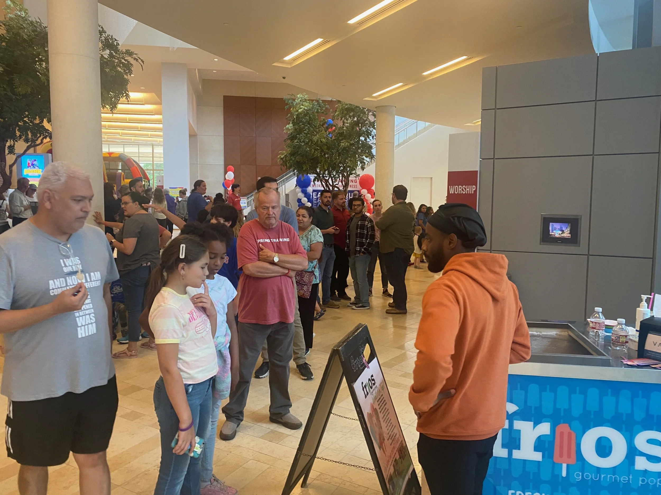 People standing in line inside a mall, with a man in an orange hoodie and black cap at a popsicle stand, surrounded by children and adults, some with crossed arms, near balloons and store signs.