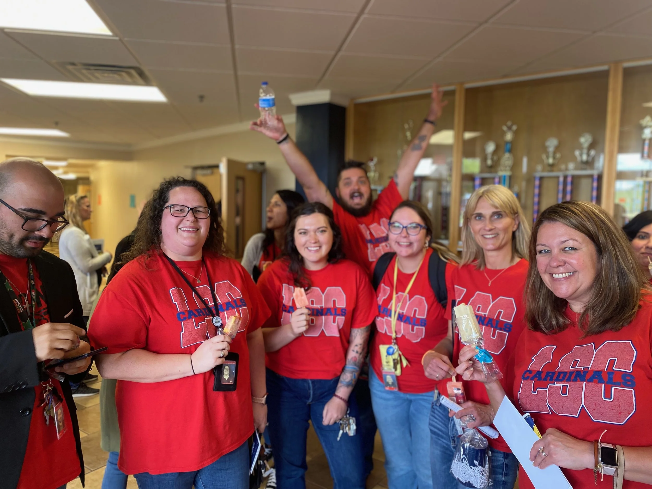 Group of people wearing red shirts with the text '100 Cardinals' celebrating indoors, some holding snacks and drinks, smiling and posing for a photo.