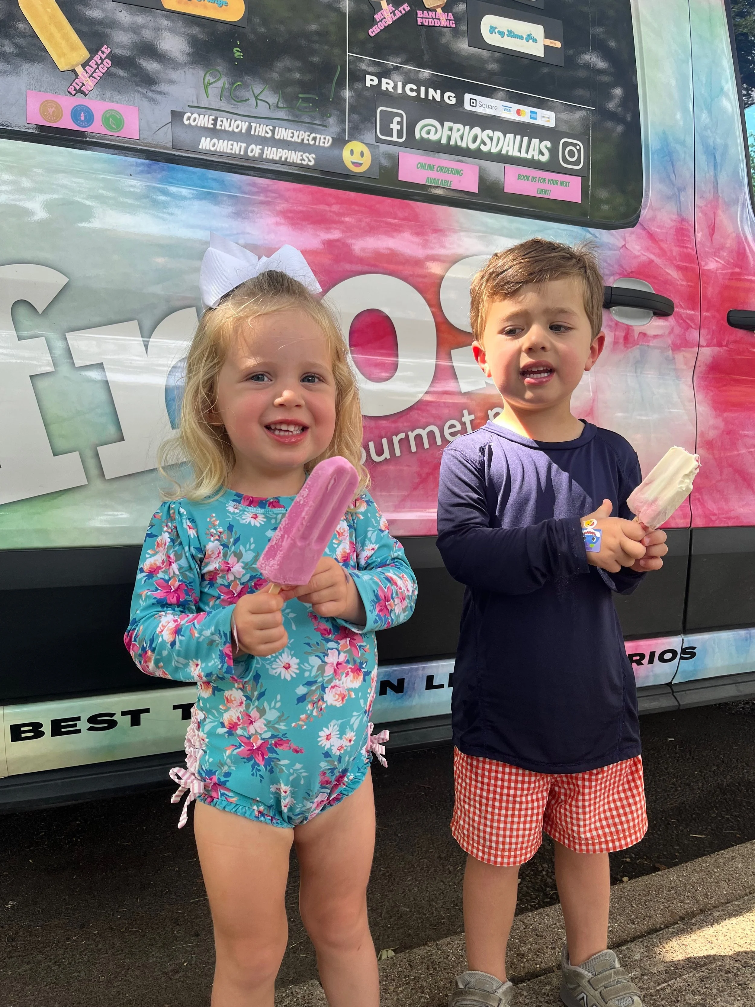 Two young children, a girl and a boy, standing in front of an ice cream truck, holding ice cream popsicles, smiling and enjoying their treats.