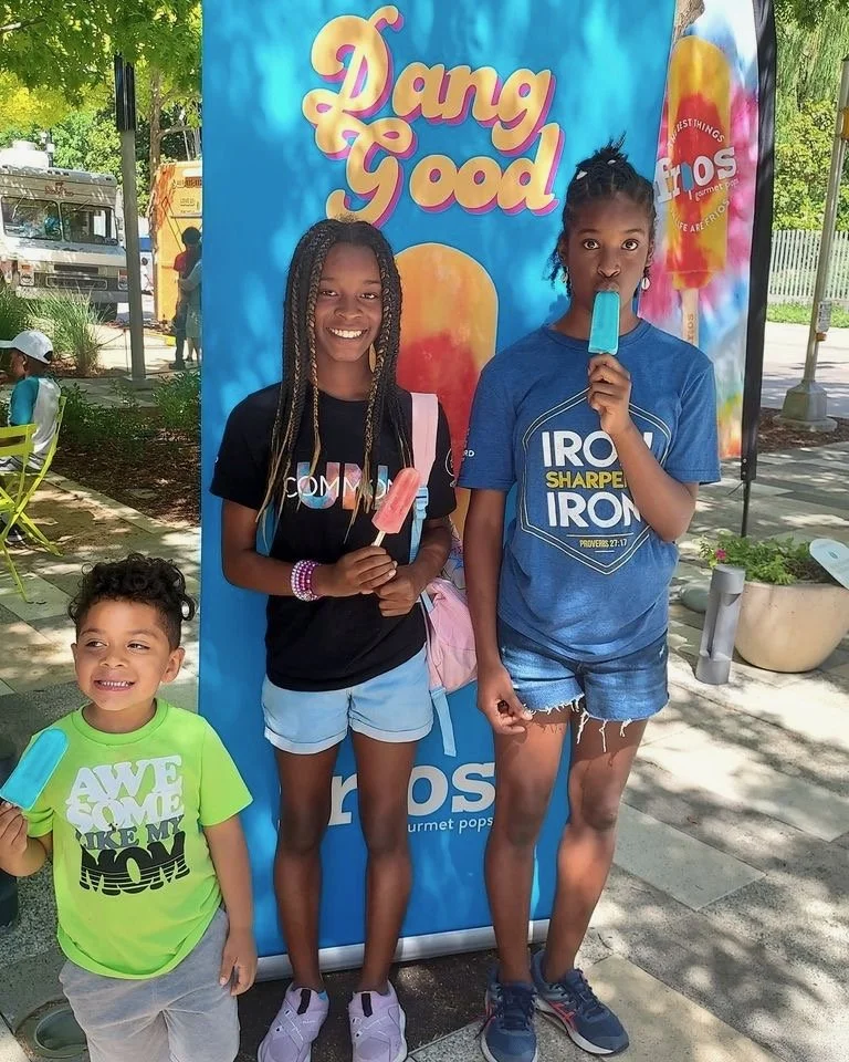 Three children standing outdoors in front of a colorful Ice Pops banner, each holding a popsicle. The youngest boy on the left wears a lime green T-shirt and gray shorts. The girl in the middle has braids, wears a black T-shirt with colorful writing,