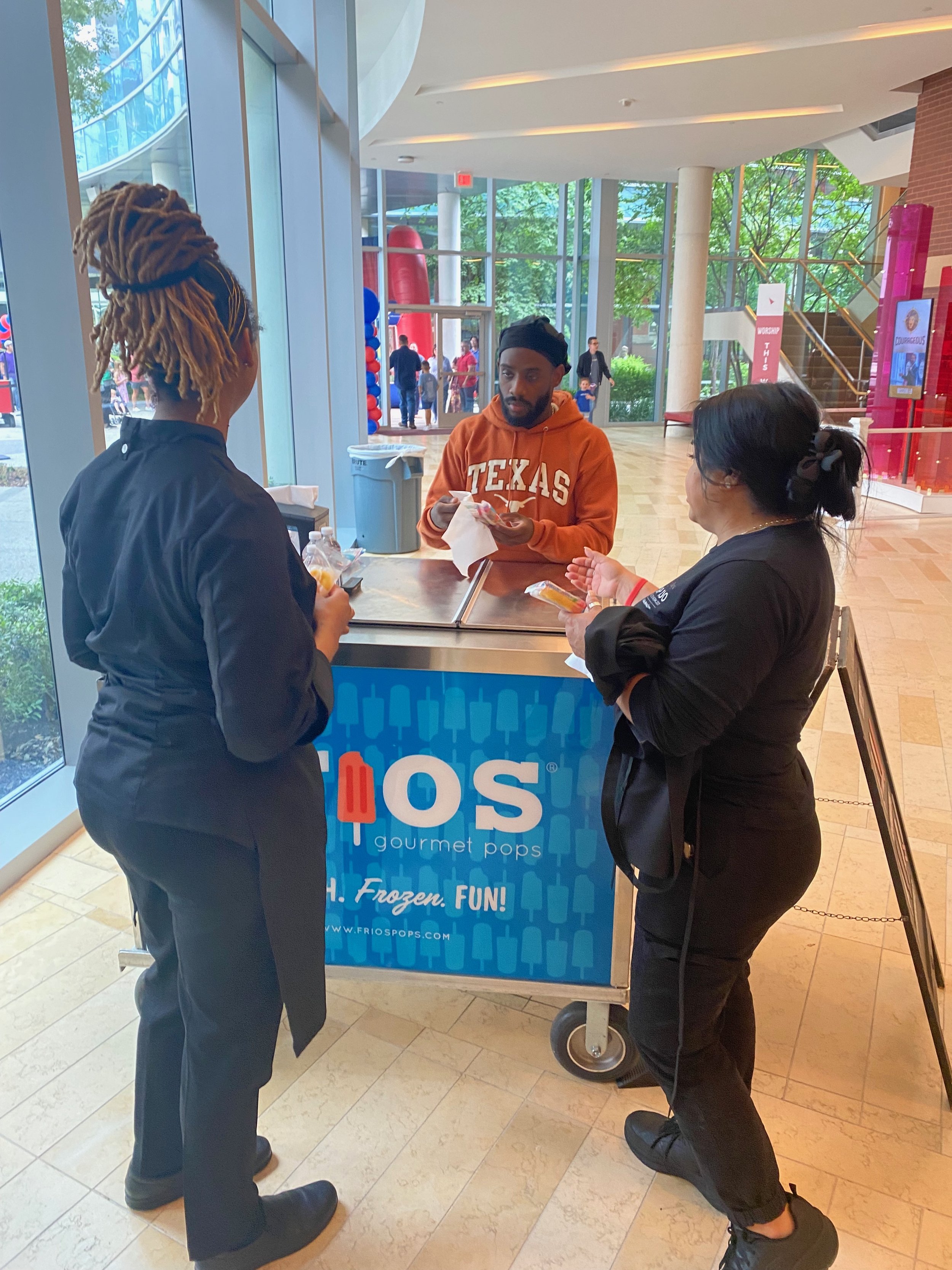 Three people standing at a frozen treat cart labeled 'frios pops' inside a building with large glass windows, next to an entrance with balloons outside.