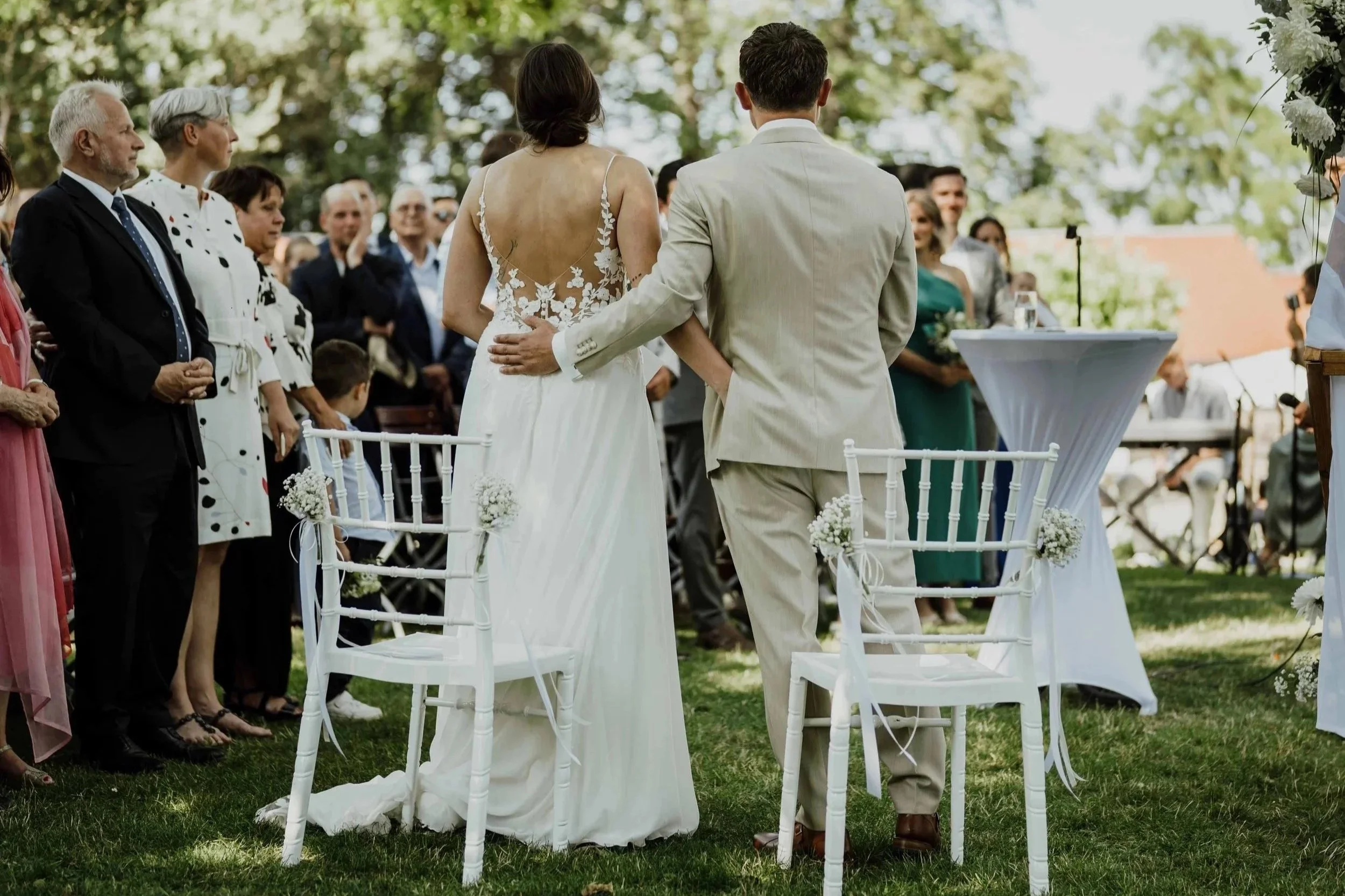 Braut und Bräutigam bei ihrer Trauung im Garten, umgeben von Gästen, mit weißen Stühlen und festlicher Dekoration.