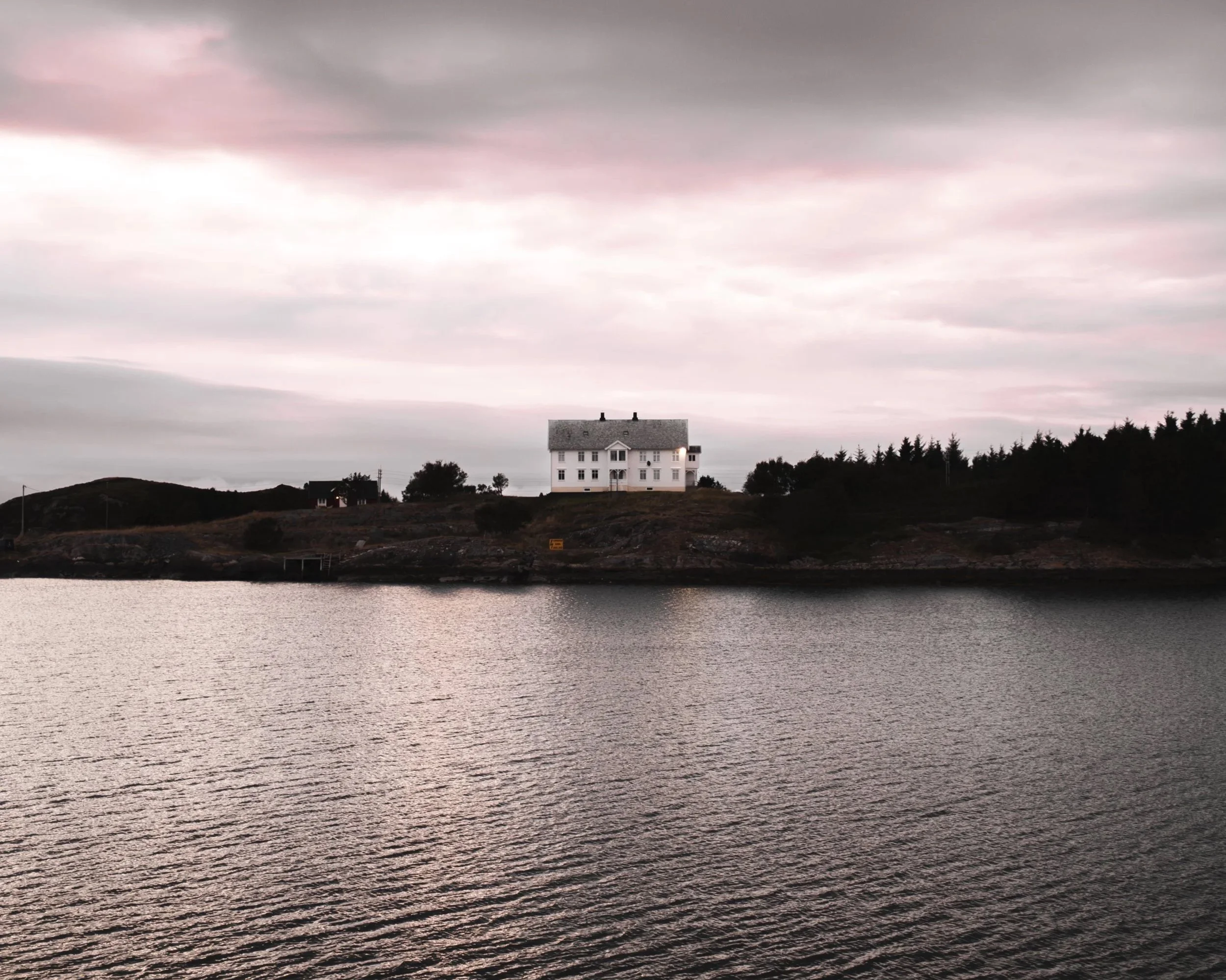 Ein Haus auf einer Insel mit Wasser im Vordergrund, bewölkter Himmel, Bäume auf einer Insel in Norwegen.