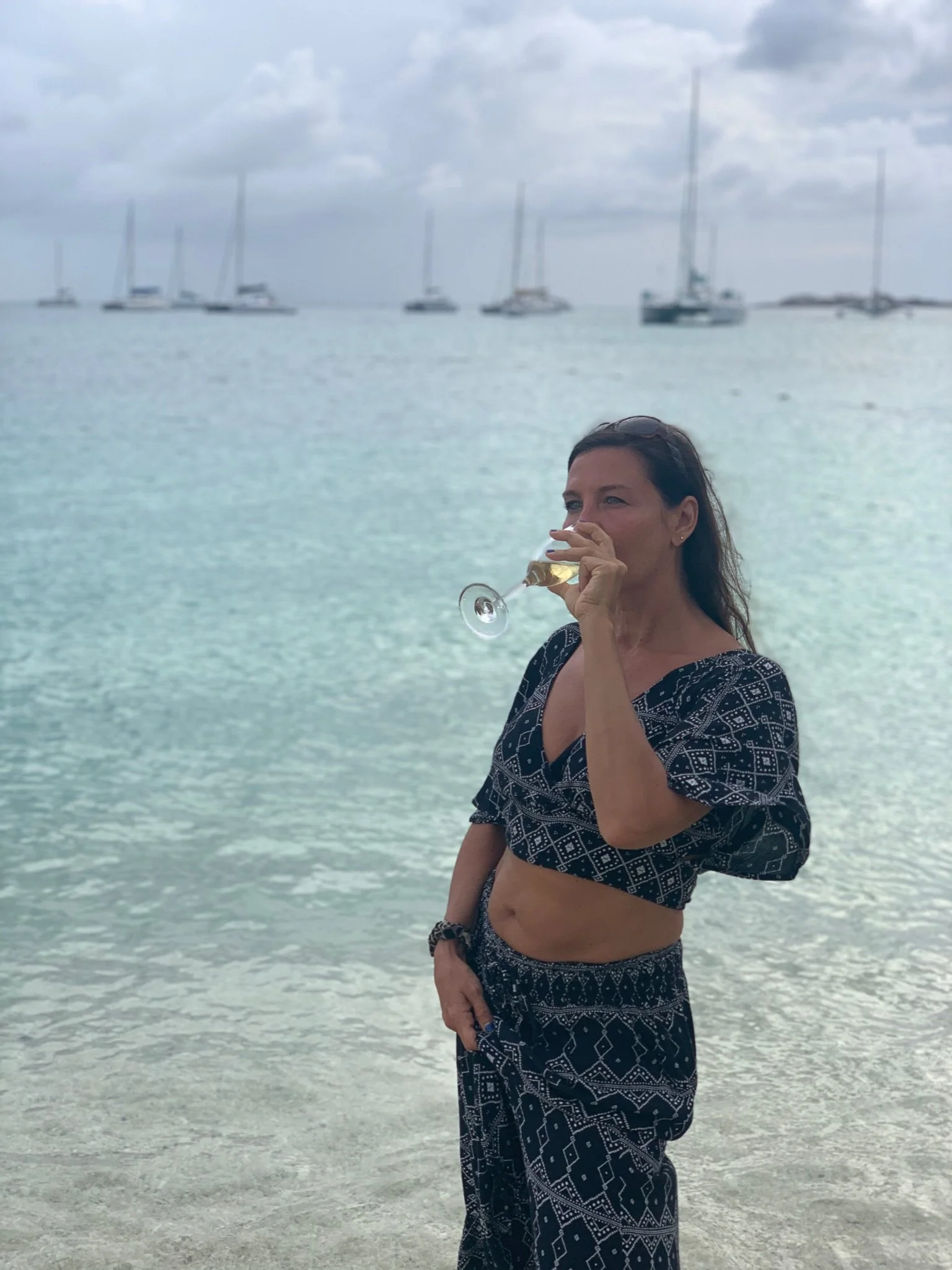Woman in a black patterned outfit standing on the beach, drinking from a wine glass, with sailboats in the water behind her and cloudy sky above.