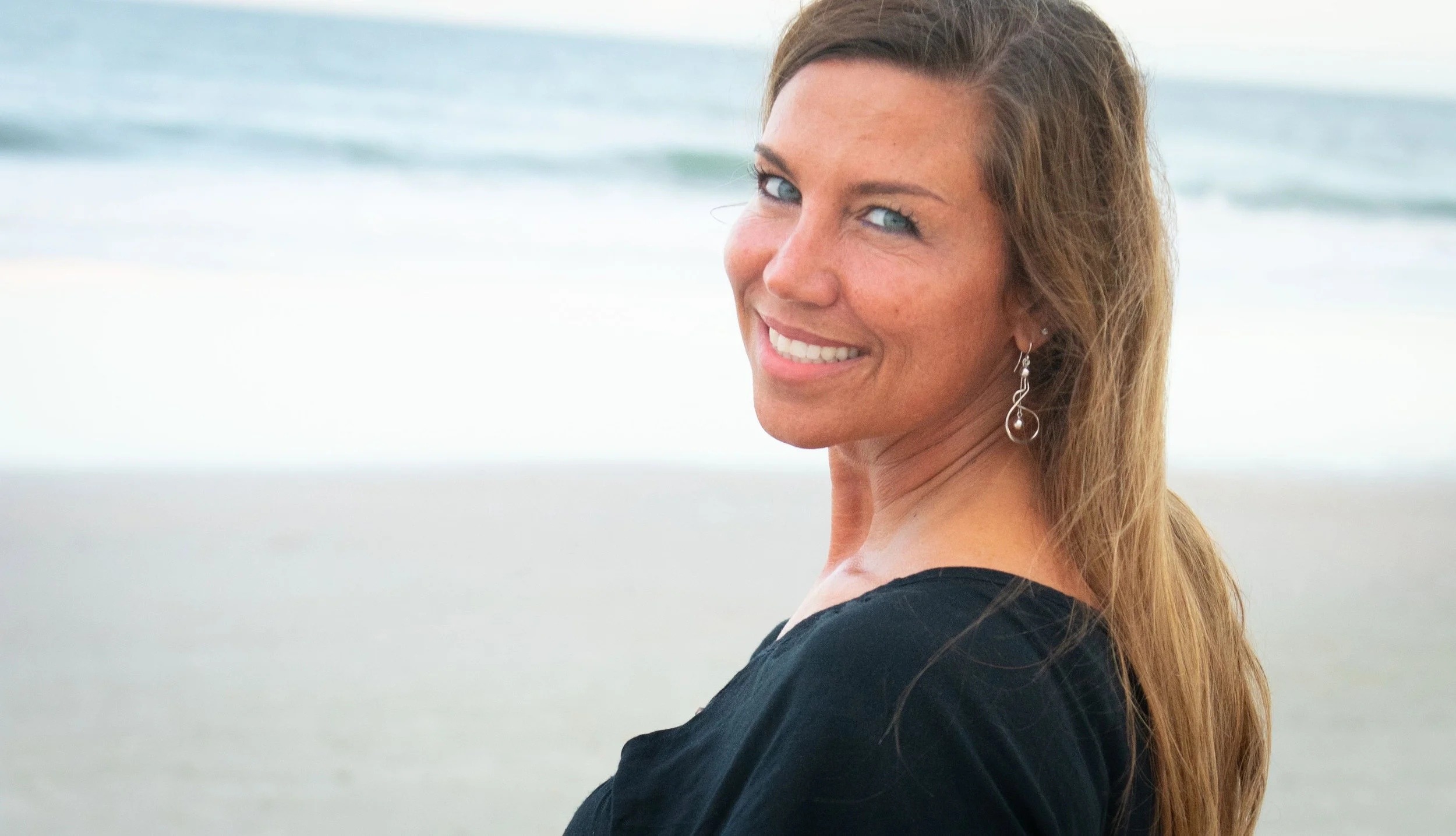 Smiling woman with long hair and earrings standing on the beach with ocean waves in the background.