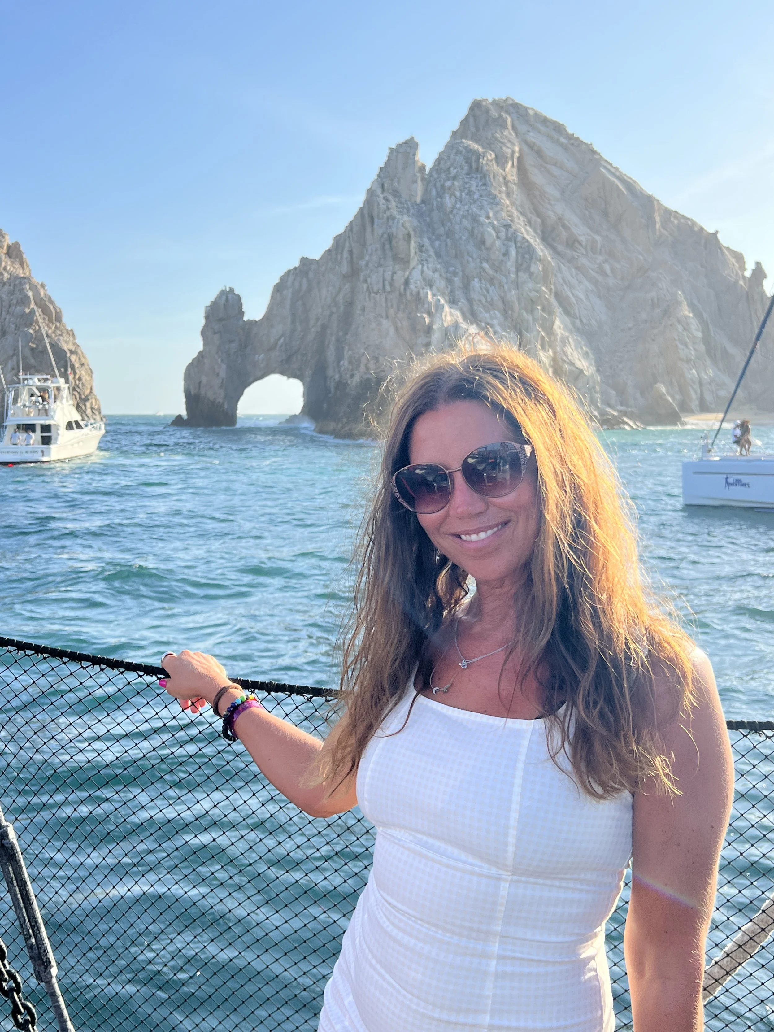 Smiling woman with sunglasses on boat near rock formation with arch in ocean