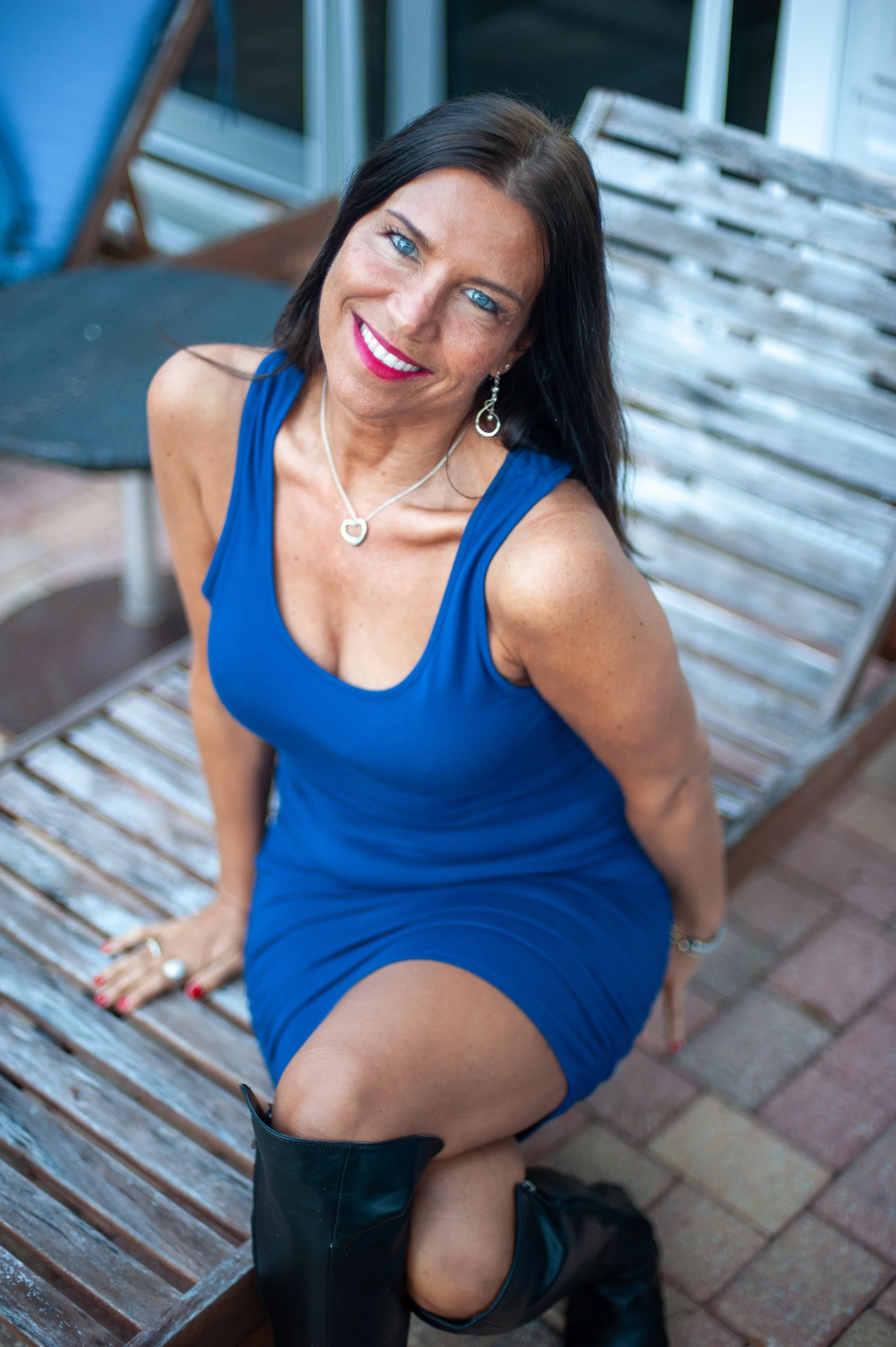A woman with dark hair and bright blue eyes sitting on a wooden bench, wearing a blue dress and black boots, smiling at the camera.