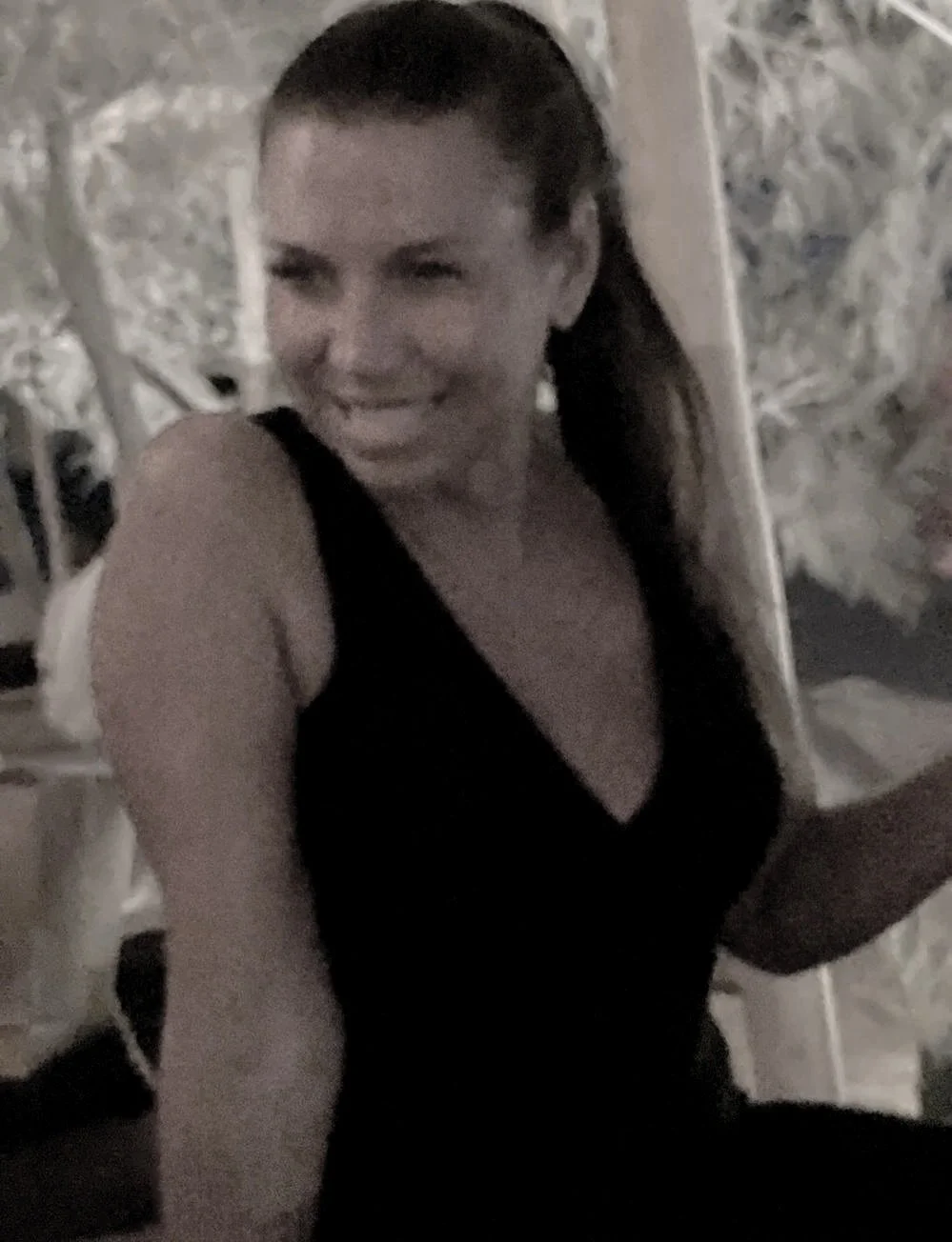 A woman with brown hair tied in a ponytail, wearing a black tank top, smiling and sitting outdoors at night.