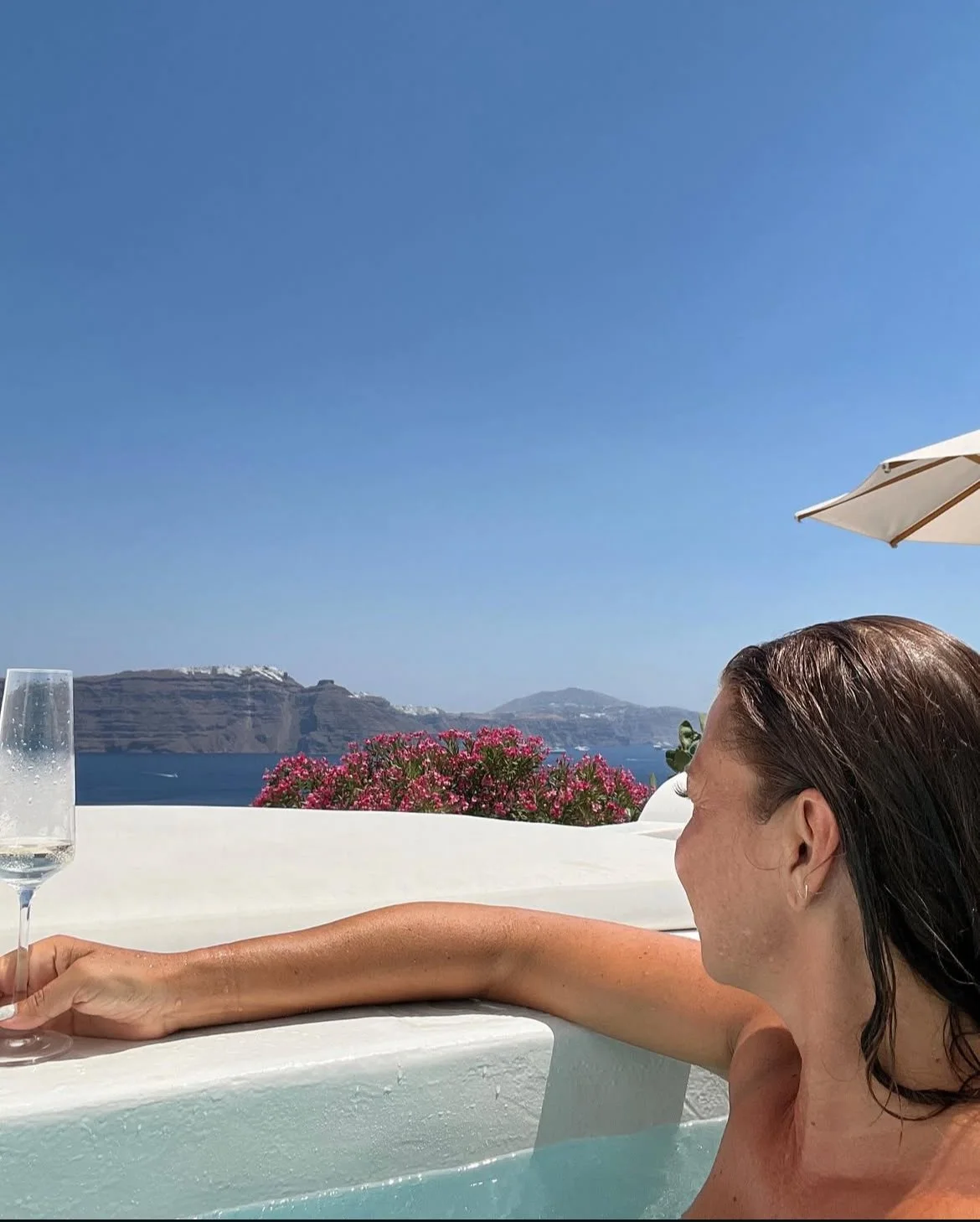 A woman relaxing in a hot tub with a view of the sea and cliffs, holding a glass of champagne, with pink flowers and blue sky in the background.